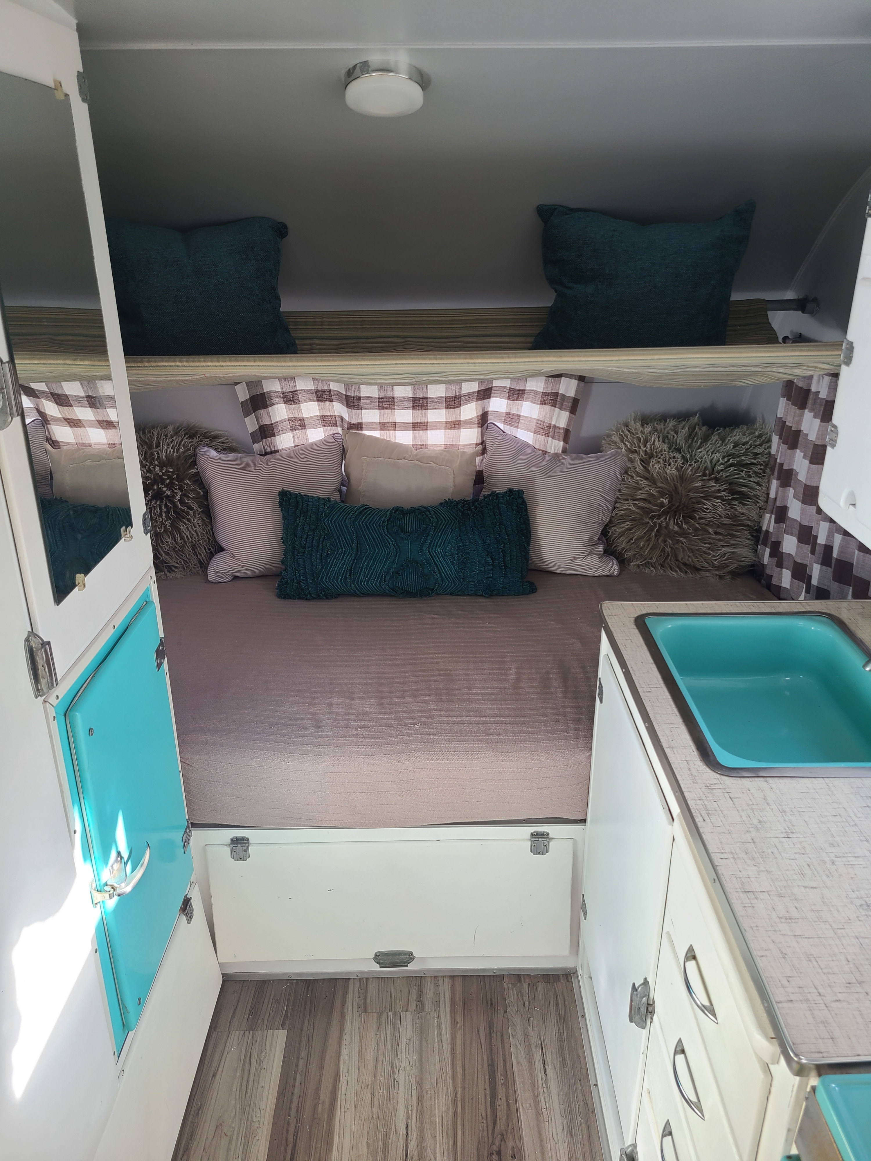 Cozy RV rental interior featuring a full bed with decorative pillows, bright teal sink, and plaid curtains.. Shasta 1500 1956