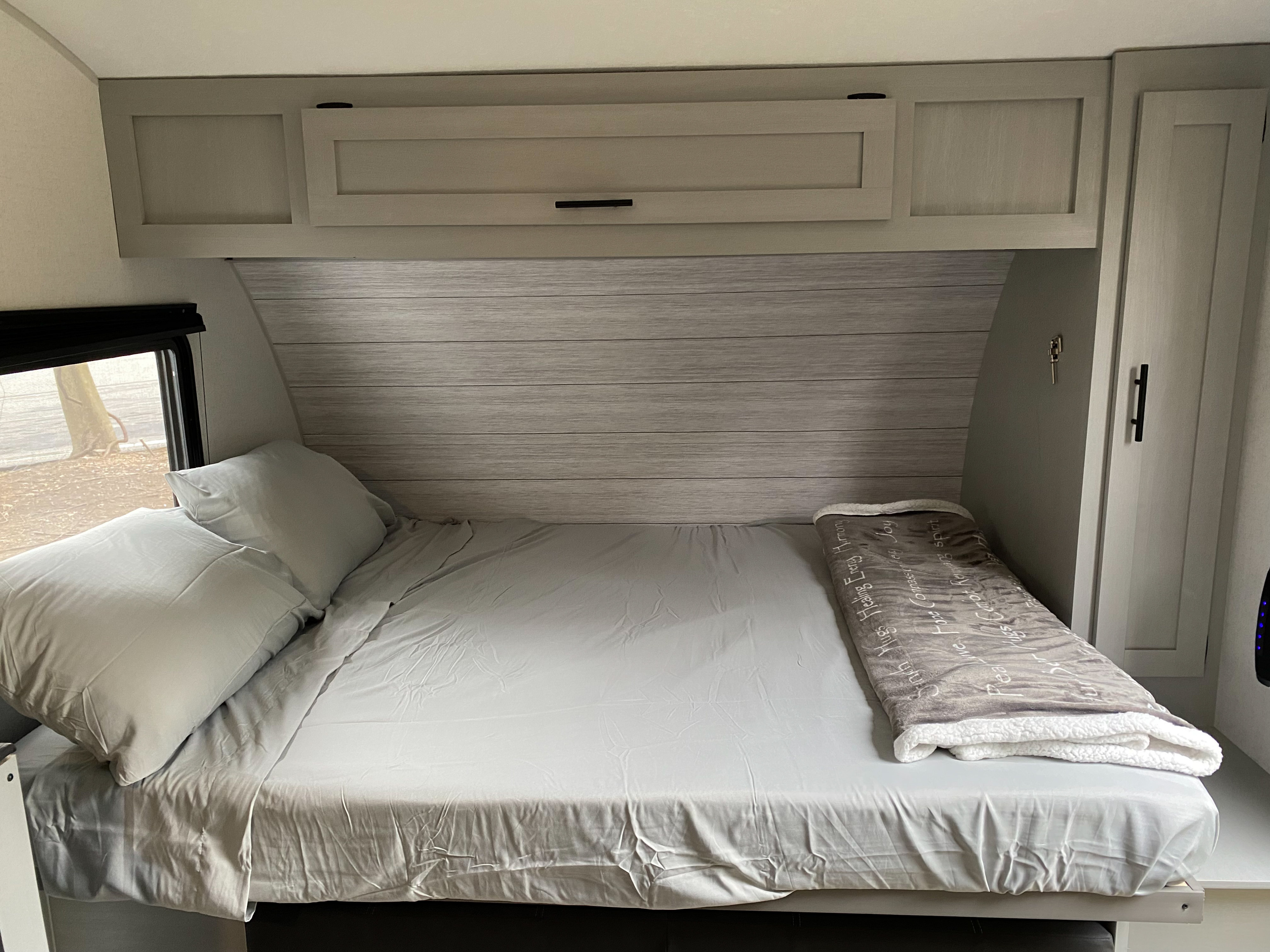 A cozy bed with light gray sheets, overhead cabinets, and a window inside an RV rental. Ideal for a comfortable sleep.. Forest River Other 2022