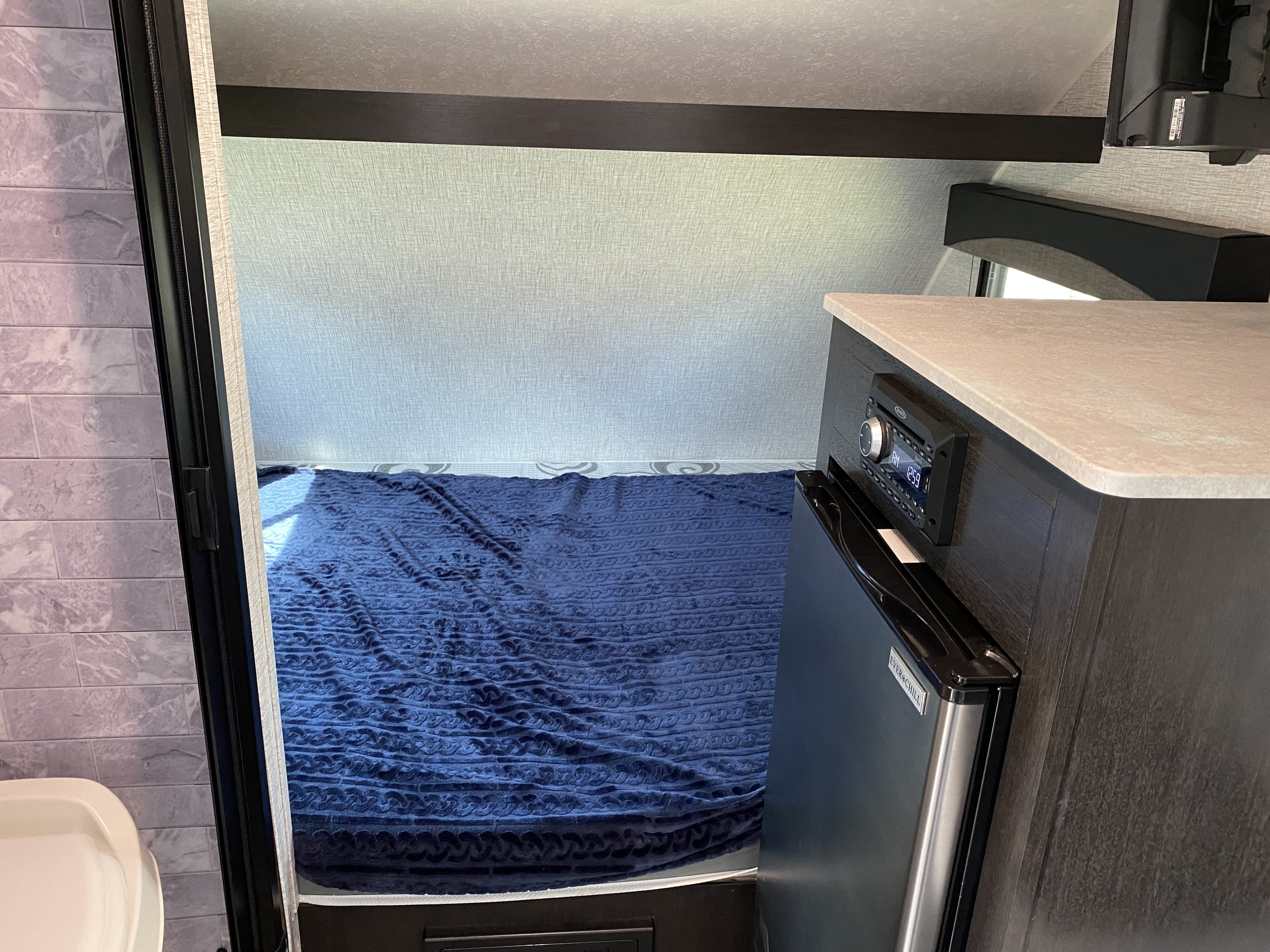 Compact RV rental interior: bed with blue blanket, kitchenette with stereo & mini-fridge, and a partial bathroom view.. Braxton Creek Bushwhacker Plus 2022