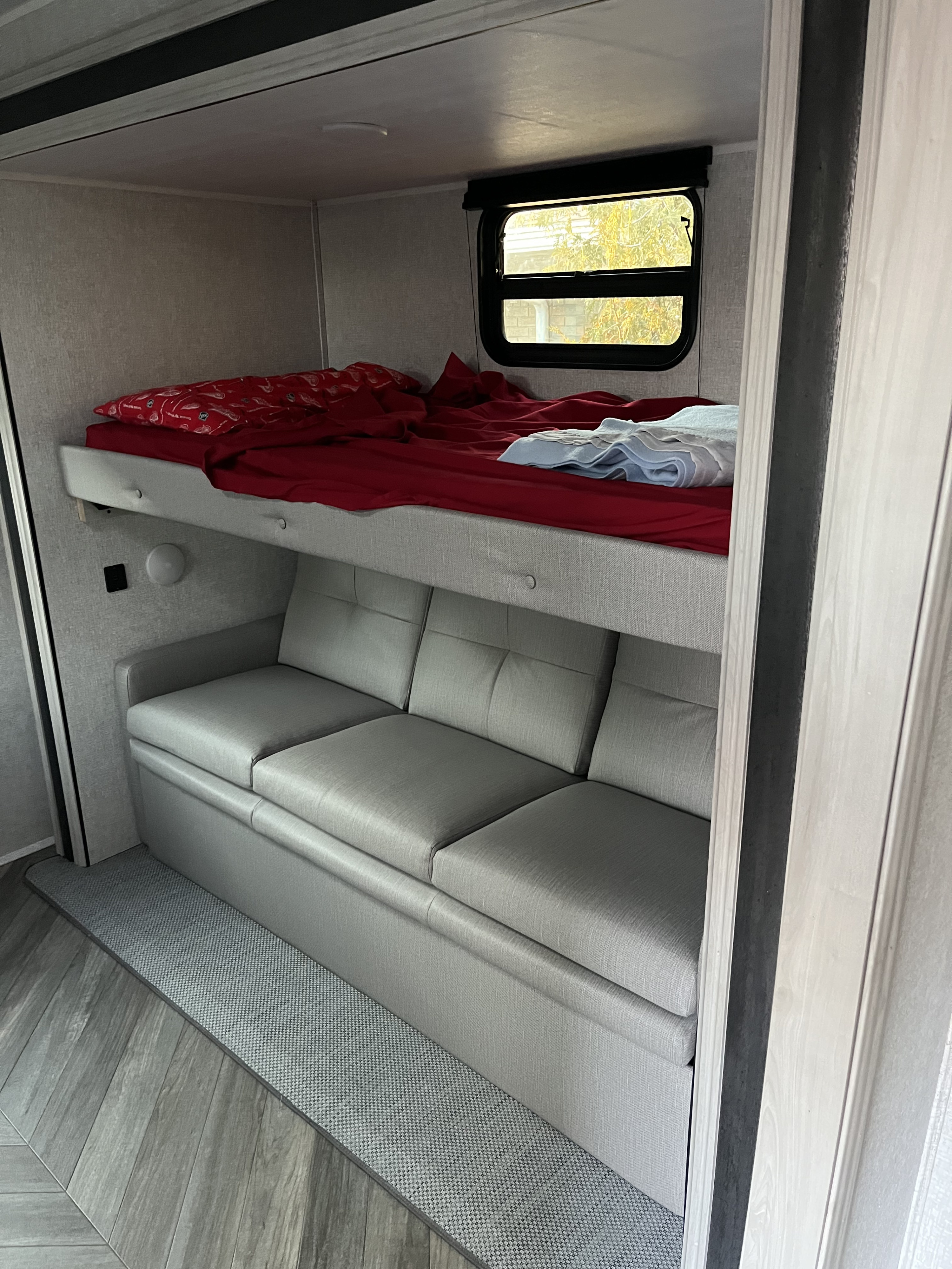 An RV rental's versatile bunk bed and sofa area. Features red bedding, a window, and gray upholstery, ideal for rest.. East to West Della Terra 2022