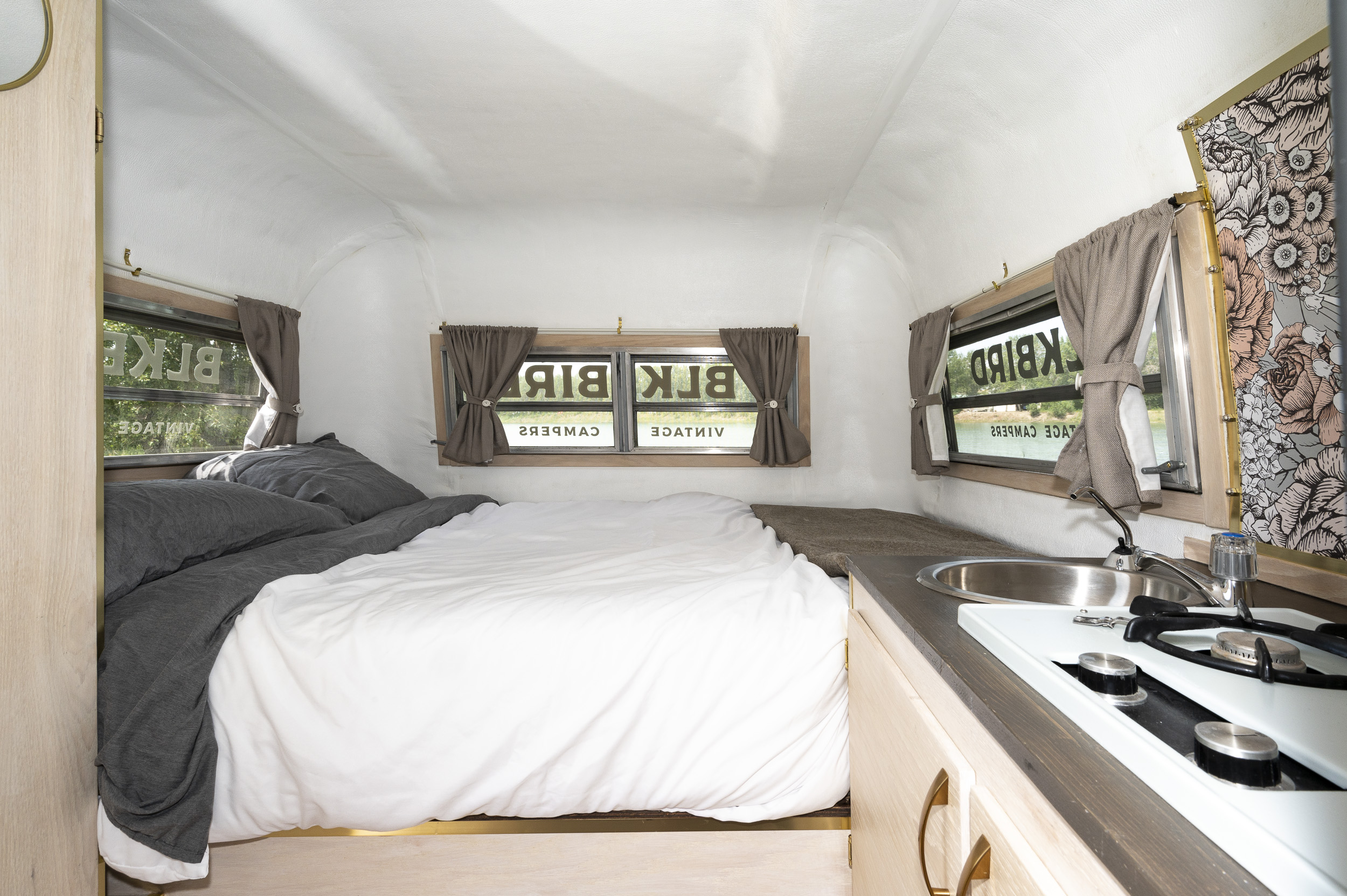 RV rental interior: cozy bed with gray and white linens, compact kitchen with sink/stove, and window views.. Triple E Surfside 1977