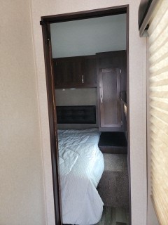 RV rental bedroom interior, seen through a doorway. Features a white bed, dark cabinets, and access to another area.. Jayco Octane 2018