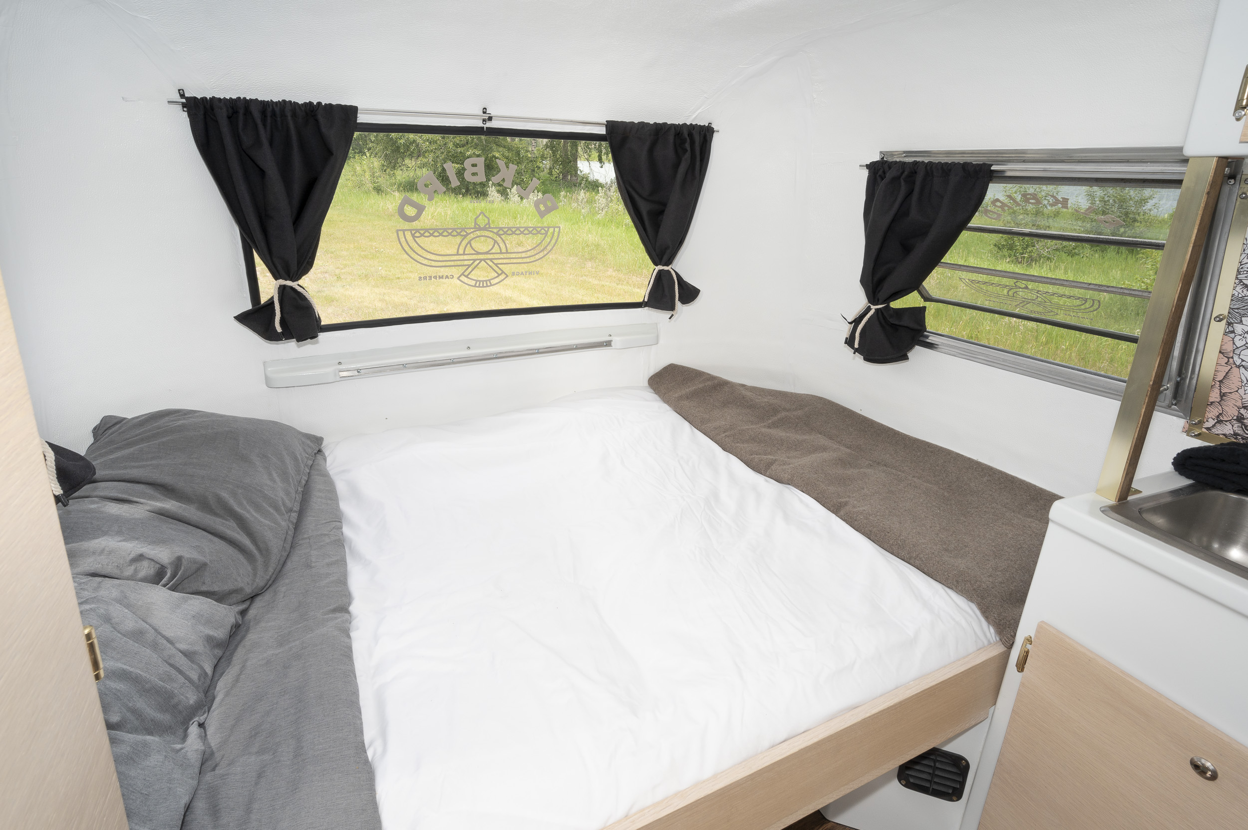 Cozy vintage rv rental interior with a bed, gray bedding, black curtains, and a compact sink, offering outdoor views.. Boler 1300 1975