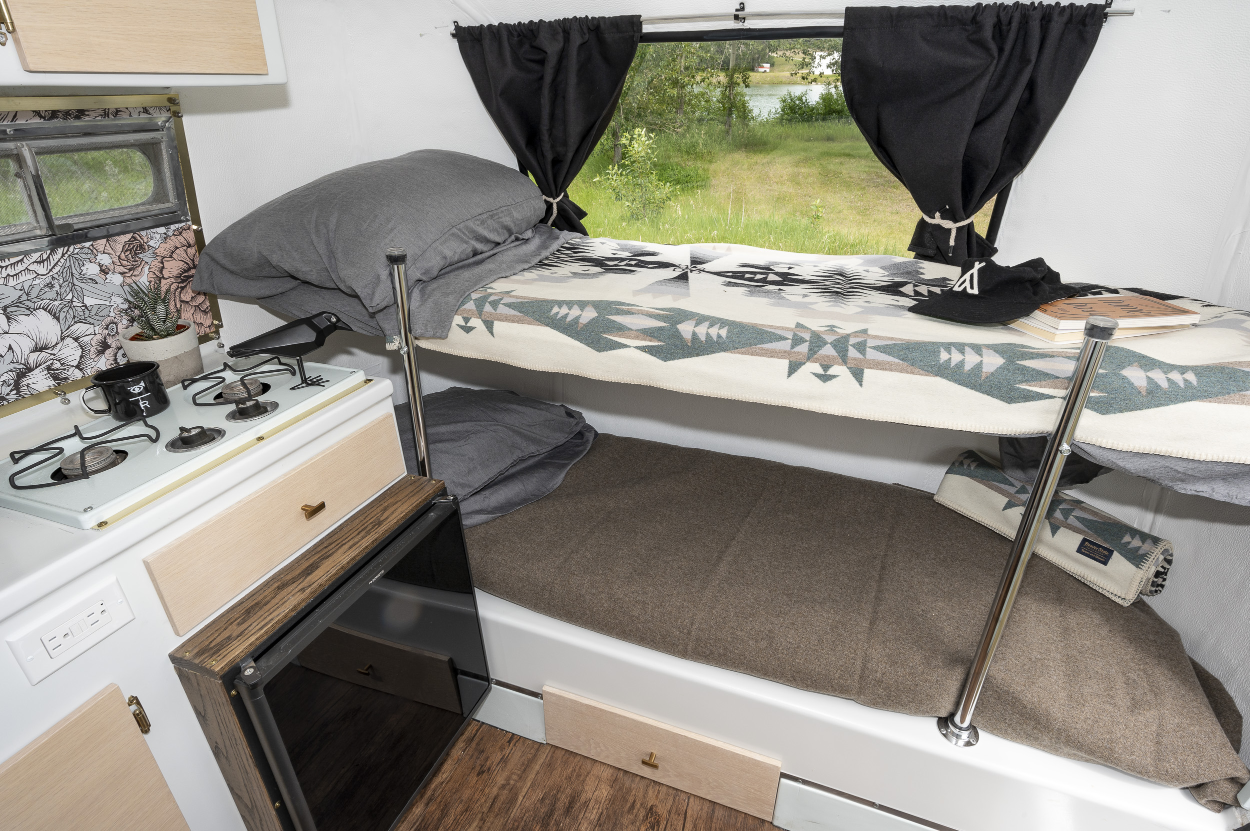 Inside a bright rv rental: bunk beds with blankets, a small kitchen area with a stove, and a window to a lake.. Boler 1300 1975