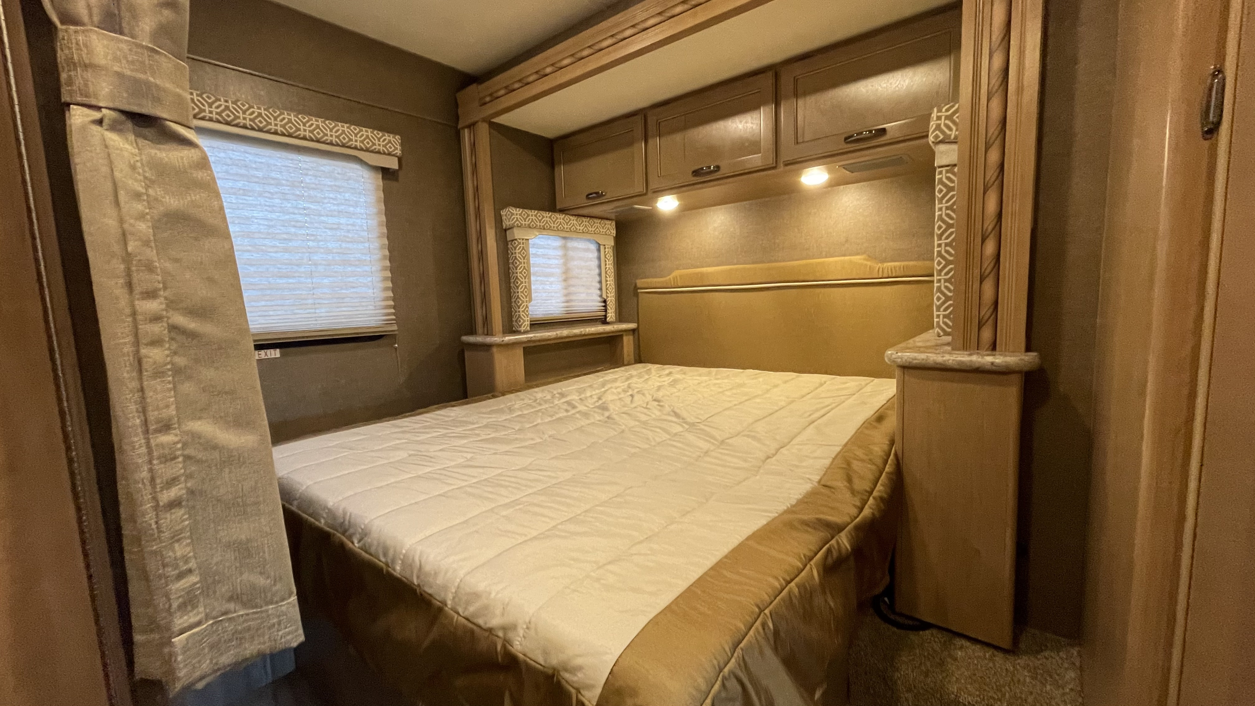 Cozy RV rental bedroom featuring a comfortable bed, overhead cabinets, and windows with patterned blinds and curtains.. Thor Chateau 2018