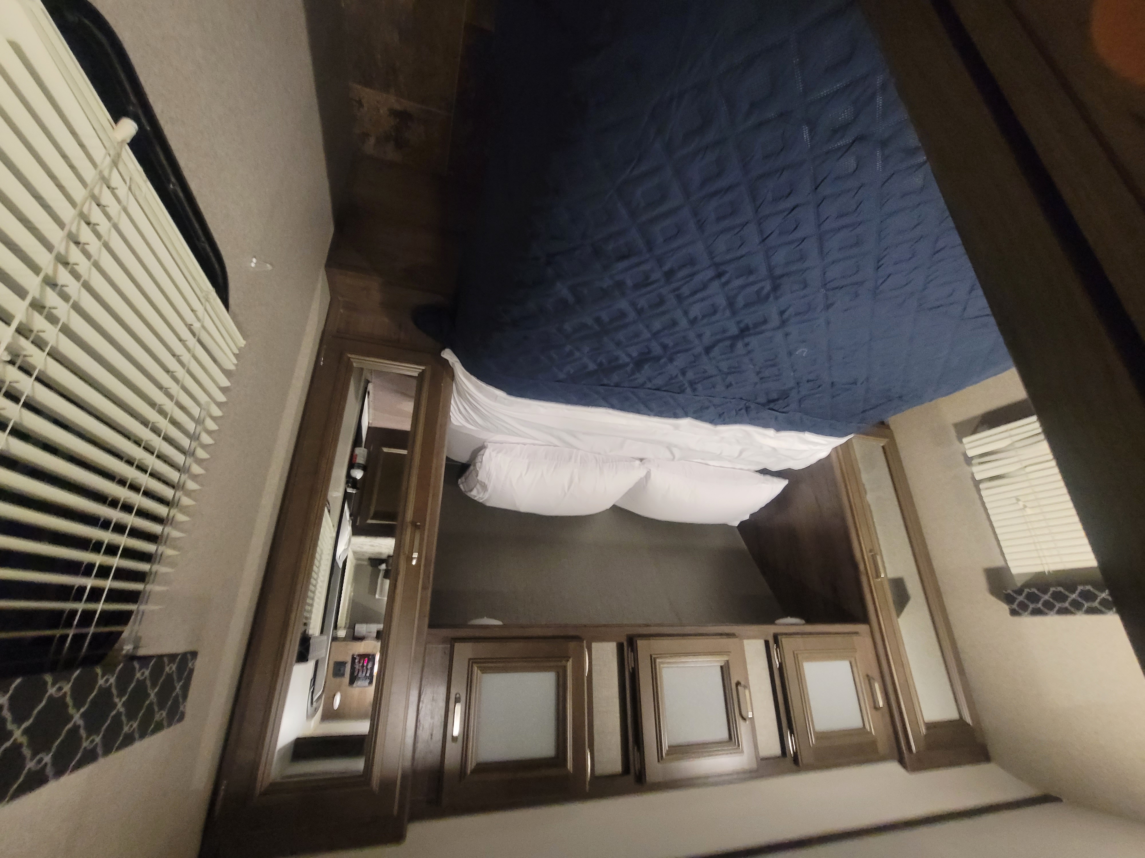 RV rental bedroom view: features a bed with blue comforter, white pillows, overhead storage cabinets, and window with blinds.. Keystone RV Passport Grand Touring 2019