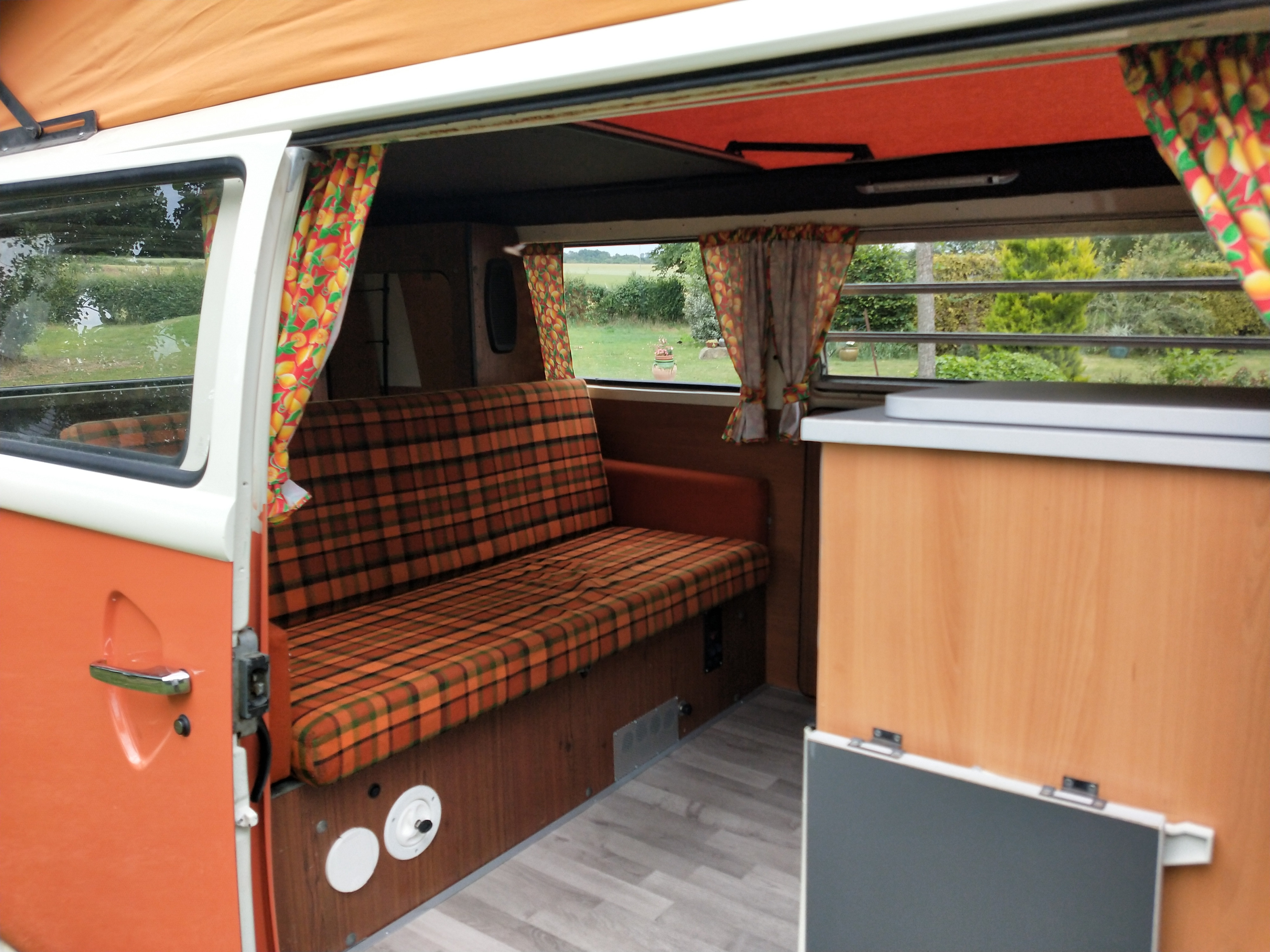 Interior of an RV rental: plaid bench seating, colorful fruit curtains, and open side door revealing a green landscape.. Volkswagen westfalia 1974