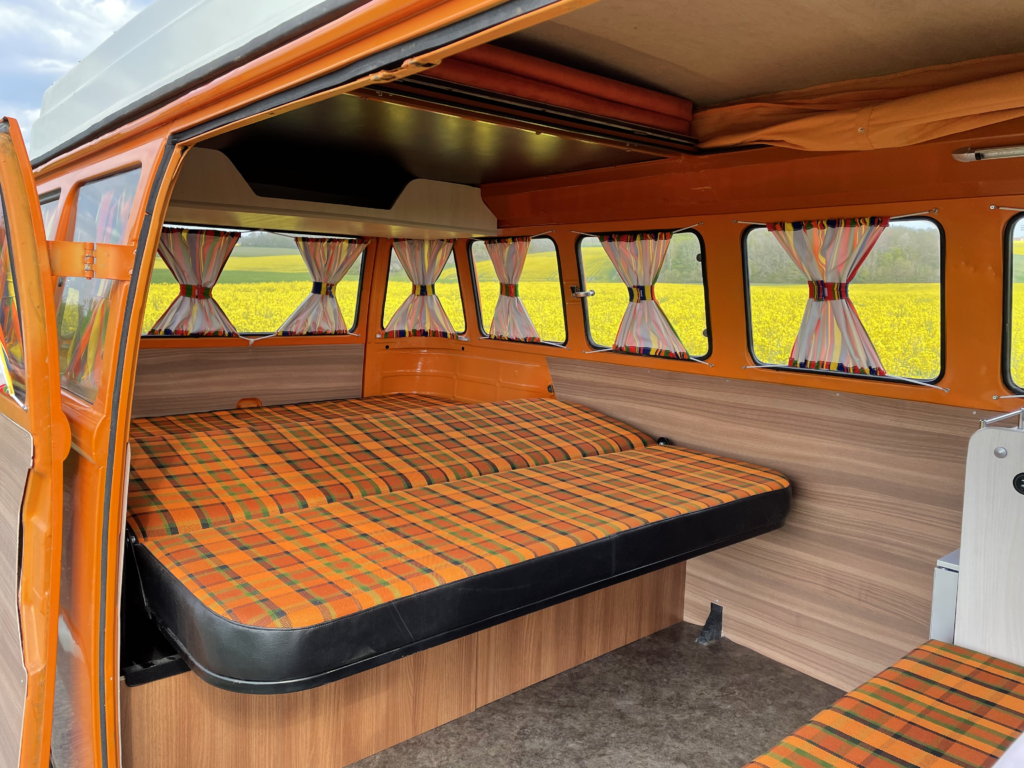 Plaid bed setup in a vintage orange camper van with colorful curtains and a bright yellow field view. Ideal for RV rental trips.. Volkswagen westfalia 1984