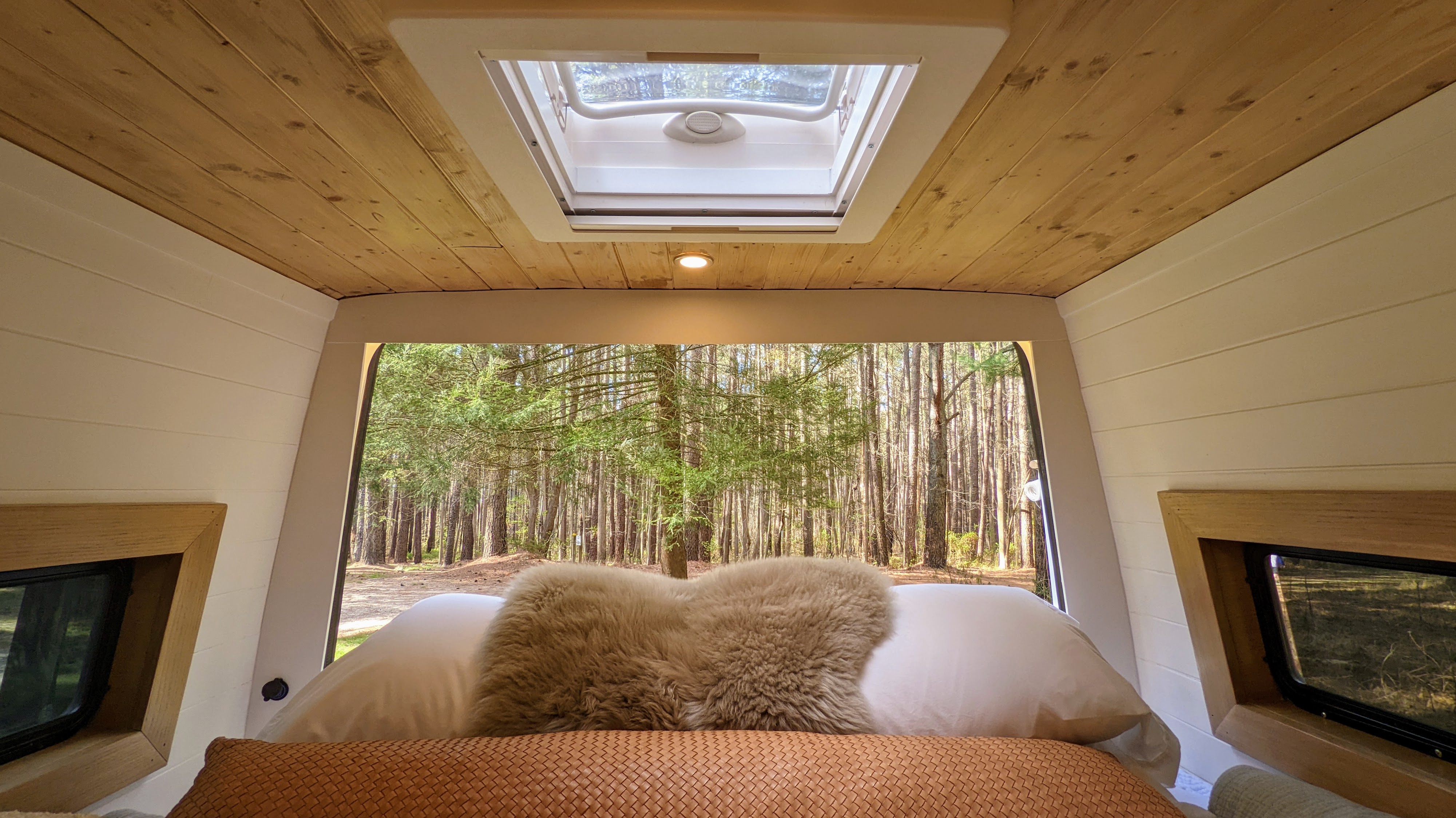 Inside an RV rental: wooden ceiling, skylight, and comfy bed facing a dense forest. Escape to nature's tranquility.. Mercedes-Benz Sprinter 2020