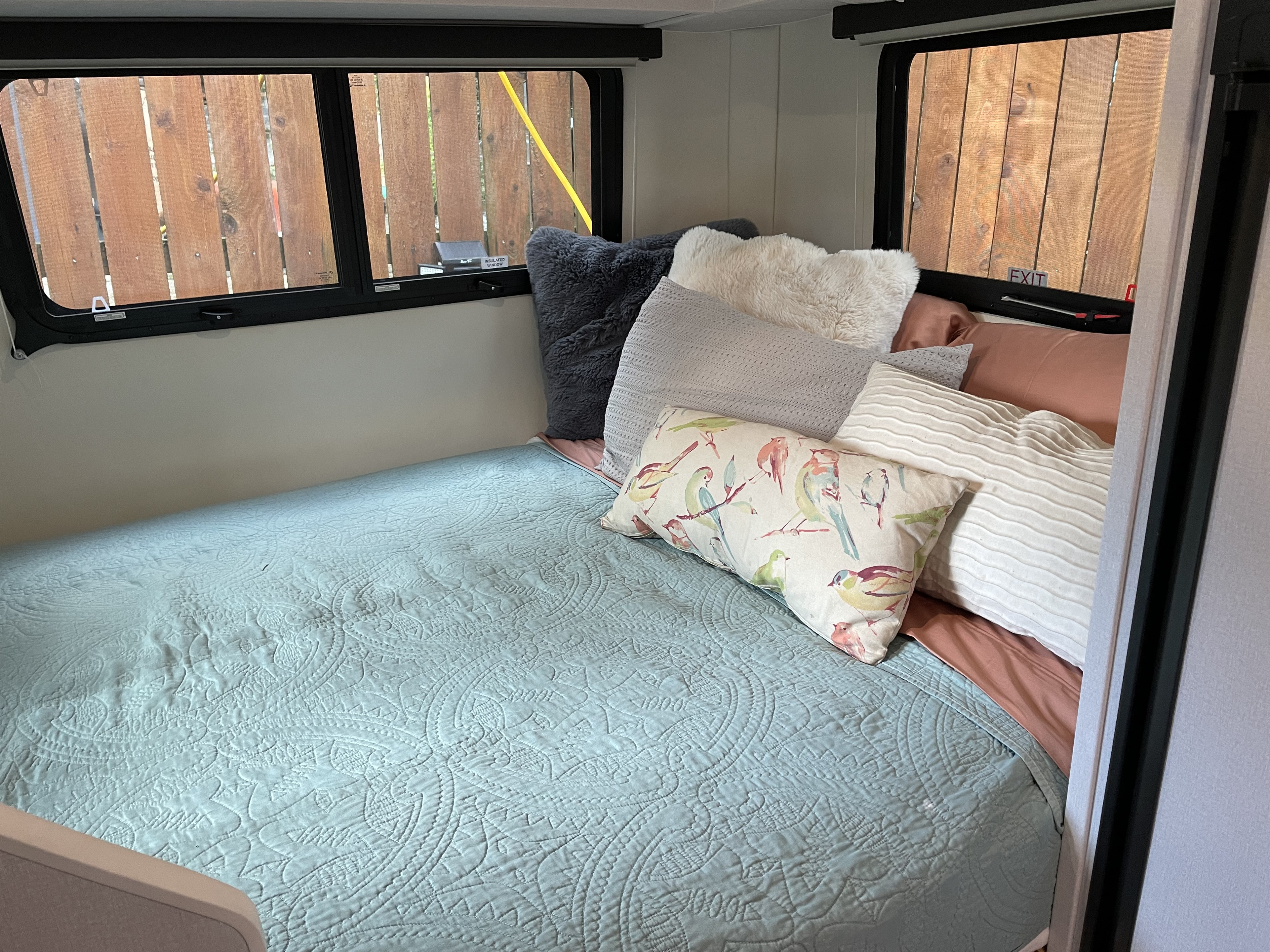 Cozy RV rental sleeping area featuring a light blue quilted bed and various decorative pillows. Windows show a wooden fence outside.. Escape E19 2022