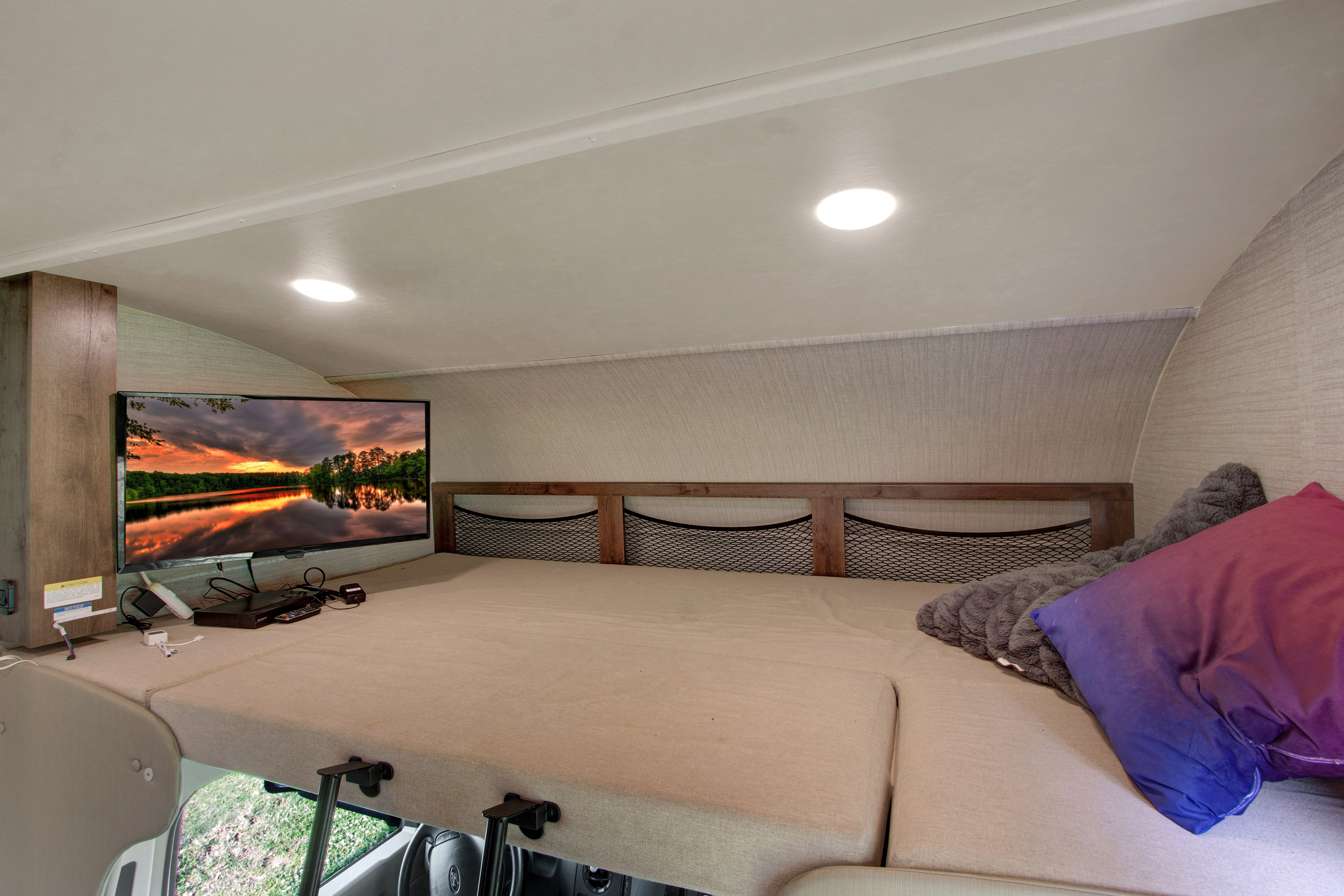 Cozy RV rental over-cab bed with TV, storage nets, and comfortable bedding, perfect for compact living on the road.. Jayco Redhawk 2020