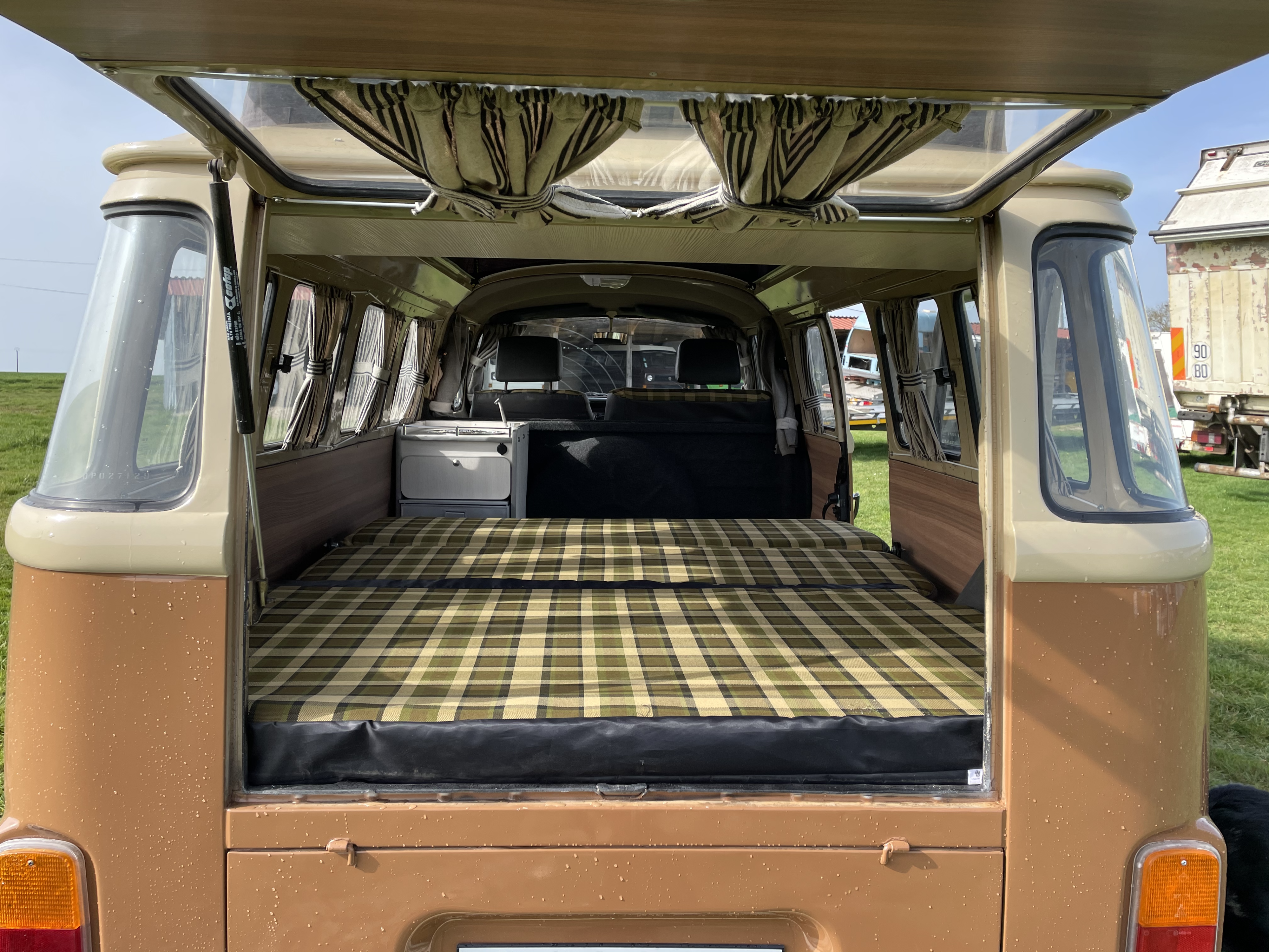 Cozy vintage camper van RV rental, showcasing a plaid bed and compact kitchen unit from its open rear. Ready for your next adventure!. Volkswagen westfalia 1983