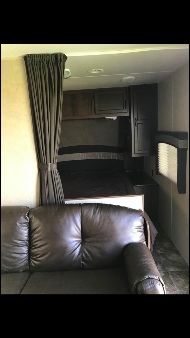 RV rental interior: A dark leather sofa and compact bedroom area with tied-back privacy curtain, bed, and cabinets.. Jayco Coyote 2014