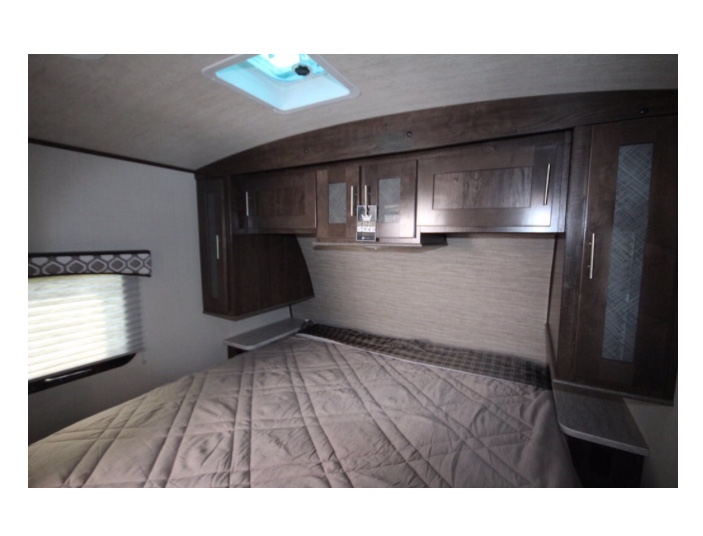 RV rental bedroom: bed with brown comforter, overhead wooden cabinets, window with blinds, and ceiling vent.. Radiance Wild Cat 2018
