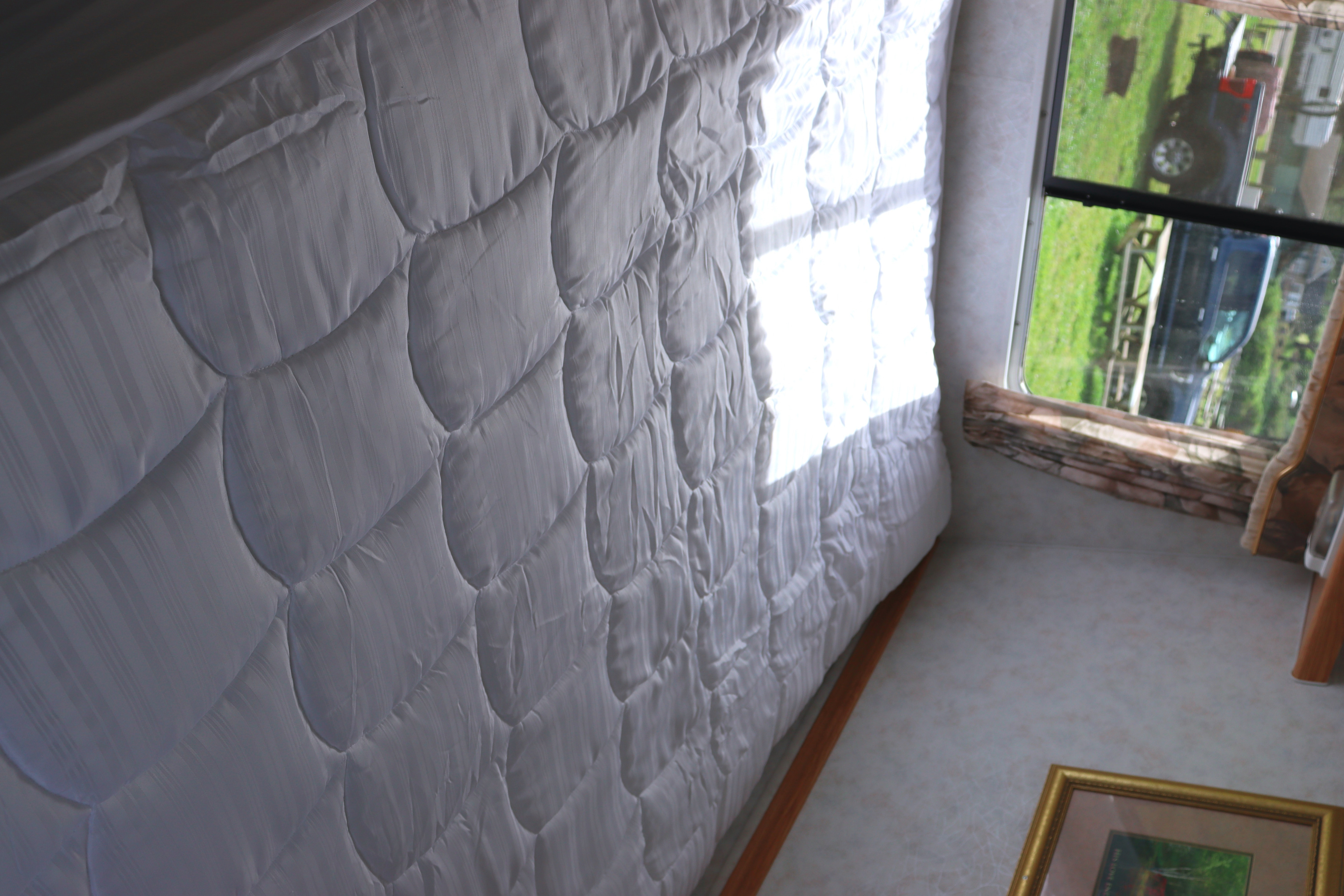 Inside an RV rental, a comfy white quilted bed is bathed in sunlight from the window, revealing a truck and green yard outside.. Komfort Komfort Trailer 2003