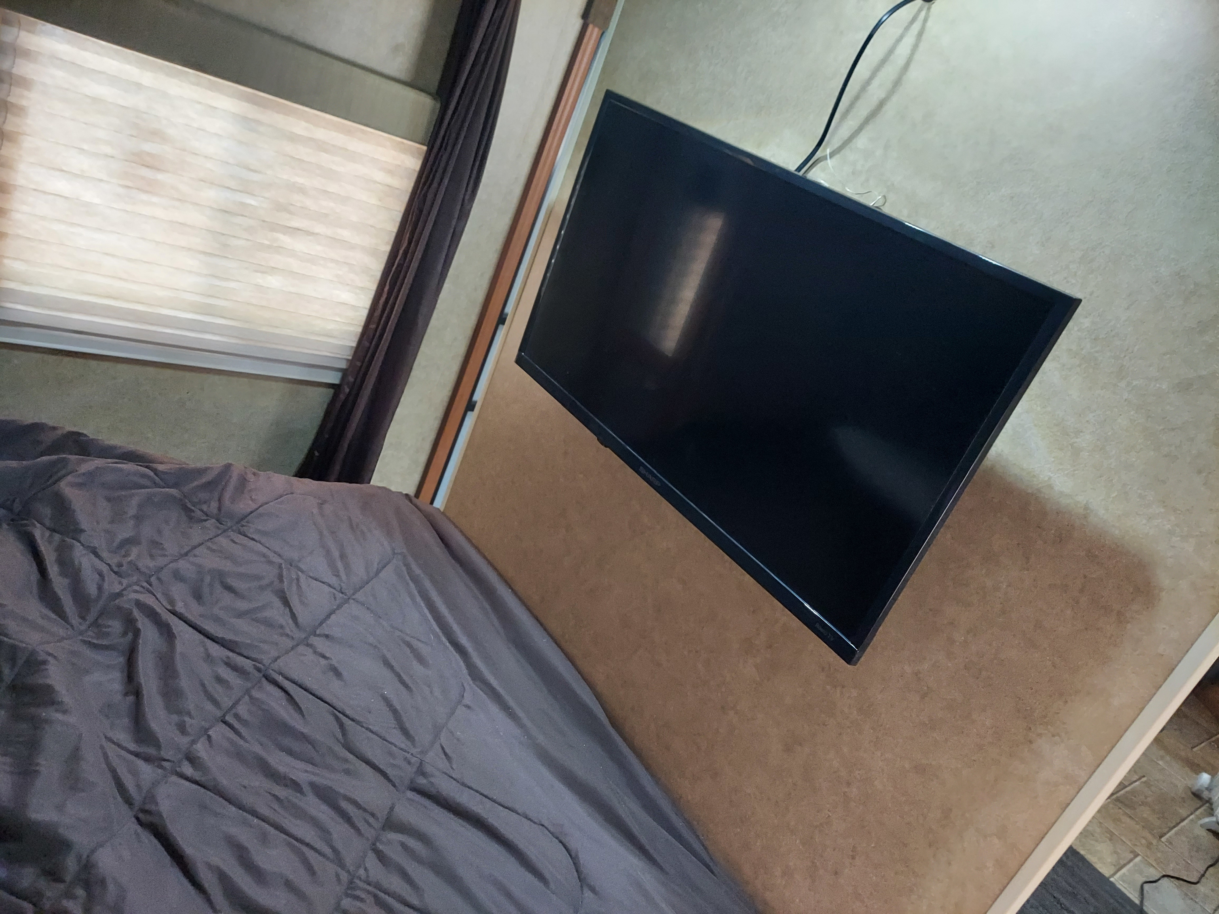Cozy RV rental bedroom showing a dark quilted bed, window with blinds, and wall-mounted TV on a brown panel.. Forest River Cherokee Grey Wolf 2014