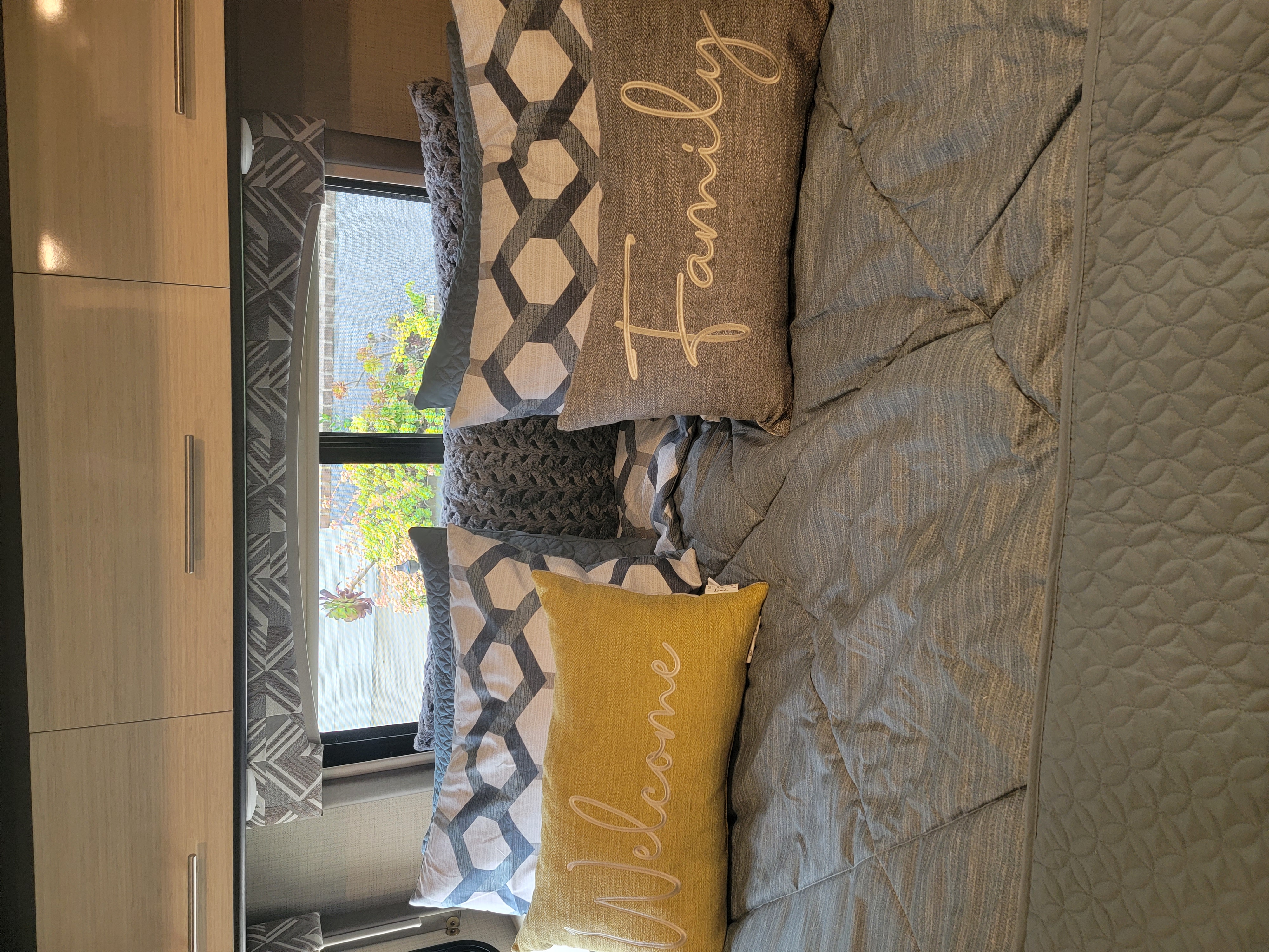 RV rental bedroom with light wood cabinets, patterned curtains, and a bed featuring "Family" and "Welcome" pillows.. Thor Axis 2021
