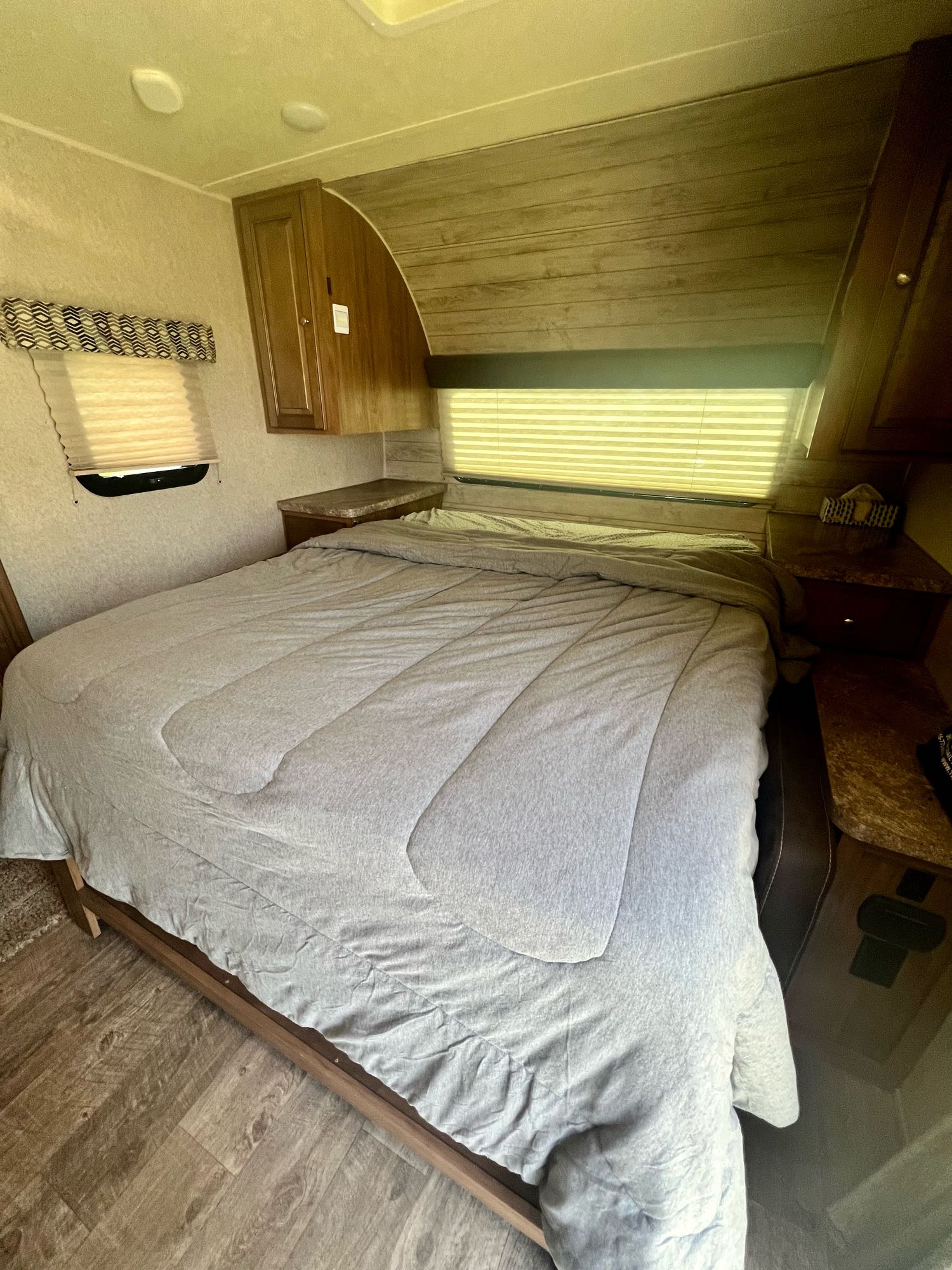 Spacious RV rental bedroom with a light gray comforter on the bed, wood floors, window blinds, and overhead storage cabinets.. Forest River Rockwood Mini Lite 2018