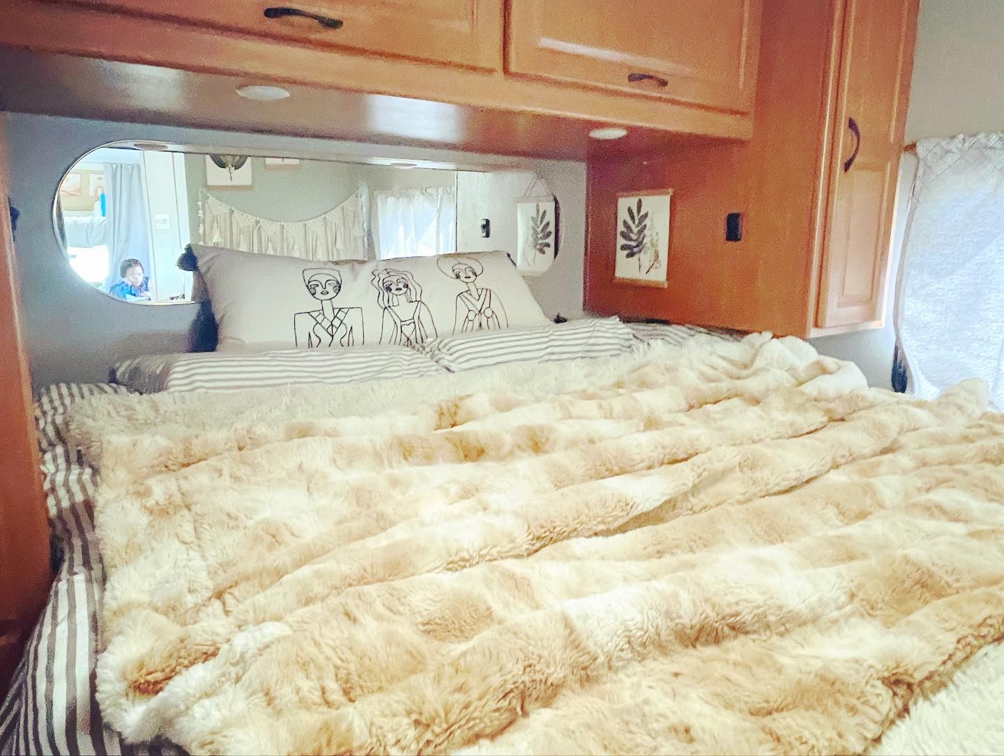 Cozy RV rental interior: soft bed with fluffy blanket, patterned pillow, and mirror reflecting a child at a table.. Thor Majestic 2018