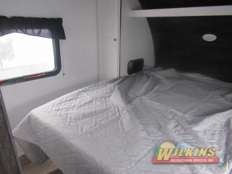 Cozy RV bedroom inside an rv rental, featuring a gray quilted bed, window with a blind, and overhead shelf.. Heartland Trailrunner 2022