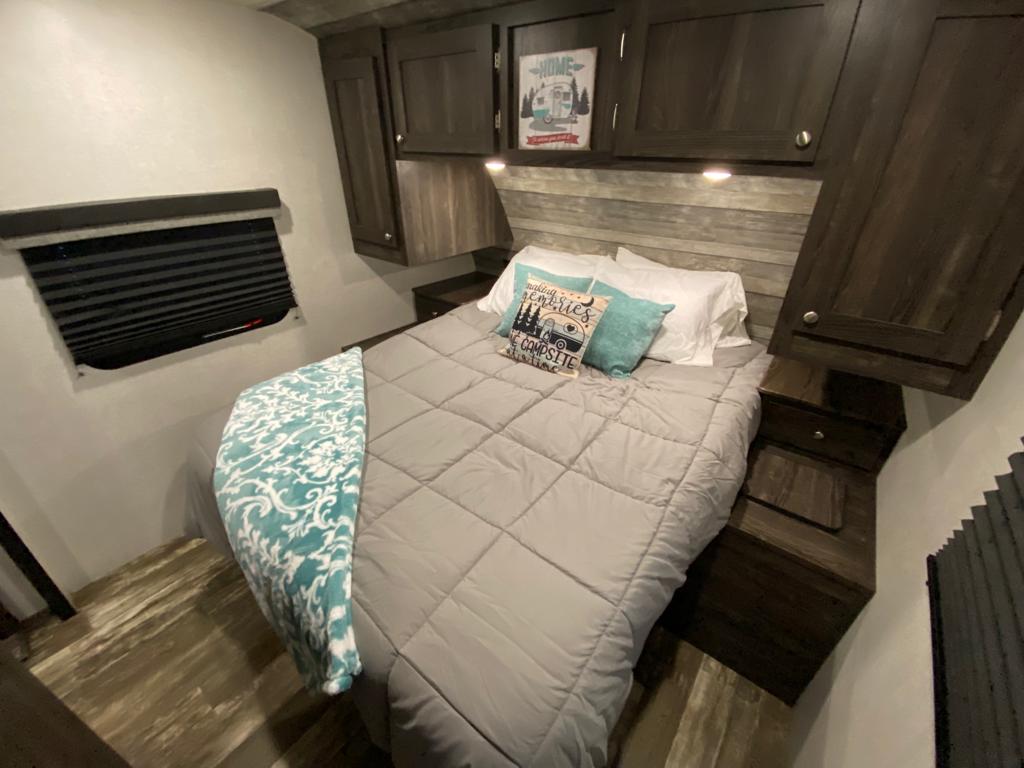 Cozy RV rental bedroom: gray comforter, teal throw, overhead storage, and window blind for travel comfort.. Starcraft Travel Trailer 2020