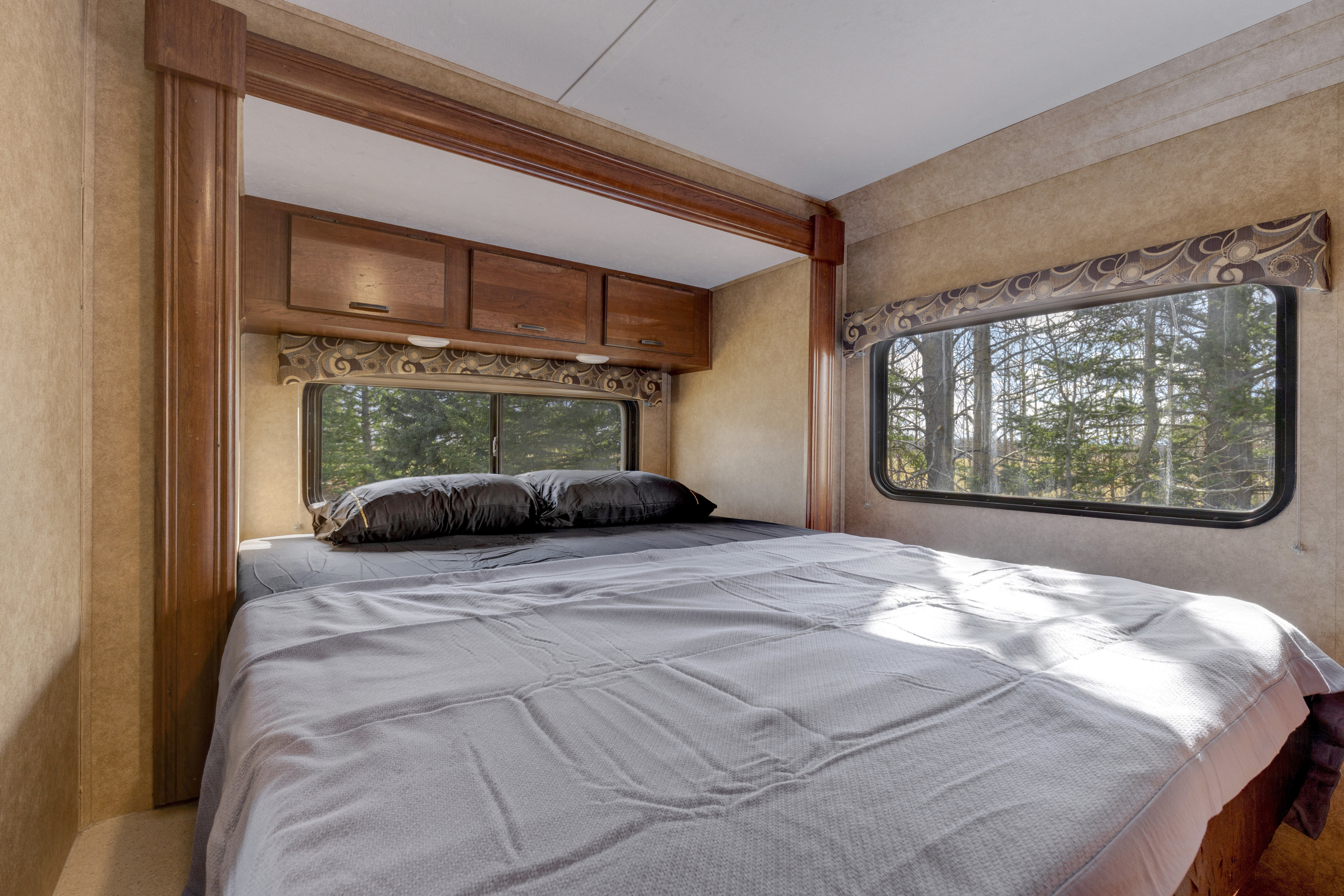 Experience this RV rental's cozy bedroom. It features a bed, overhead storage, and windows framing a sunlit forest view.. Forest River Sunseeker 2014