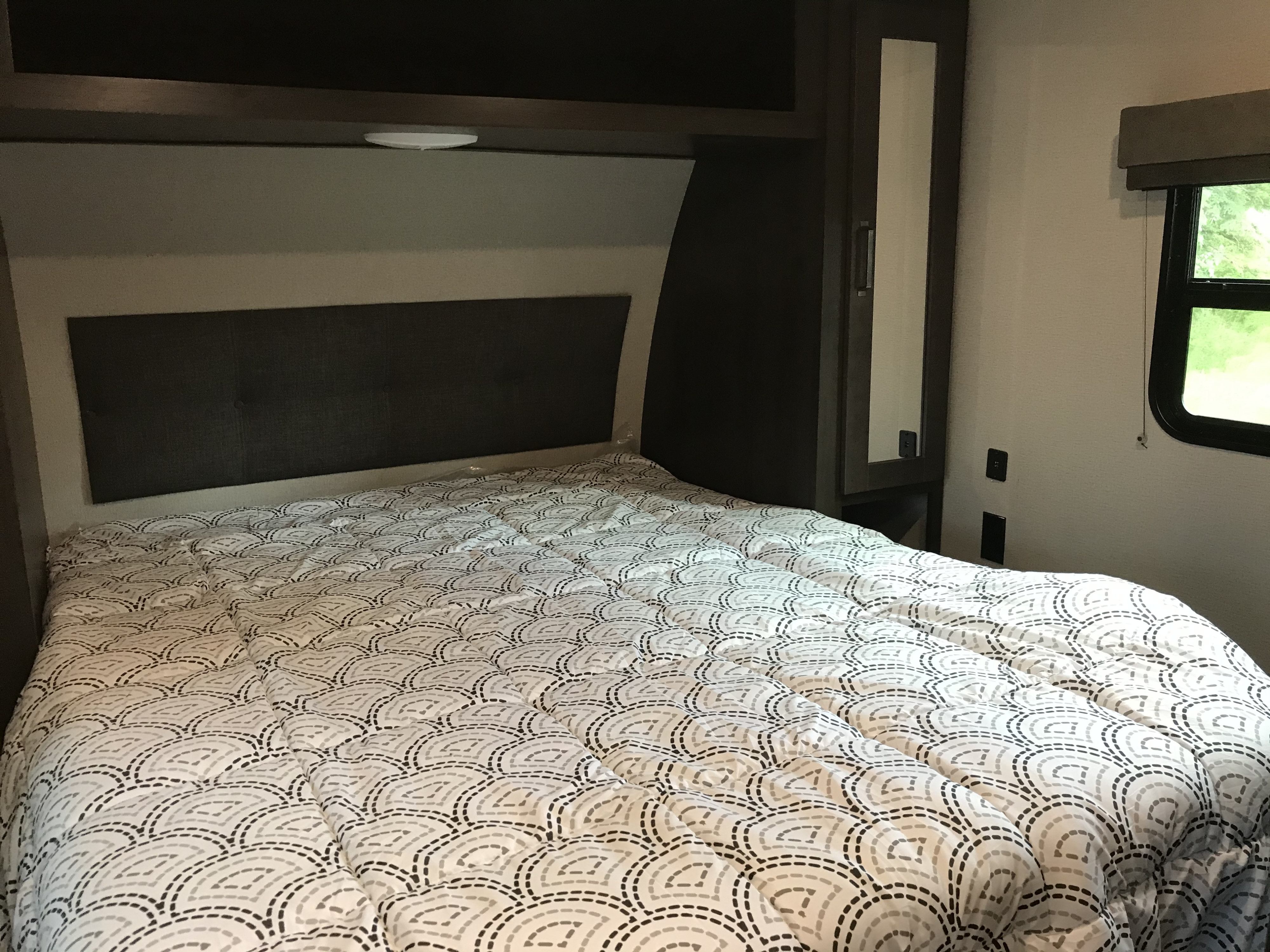 Comfortable RV rental bedroom featuring a bed with a patterned comforter, dark built-in cabinetry, and a window.. Grand Design Transcend Xplor 2021