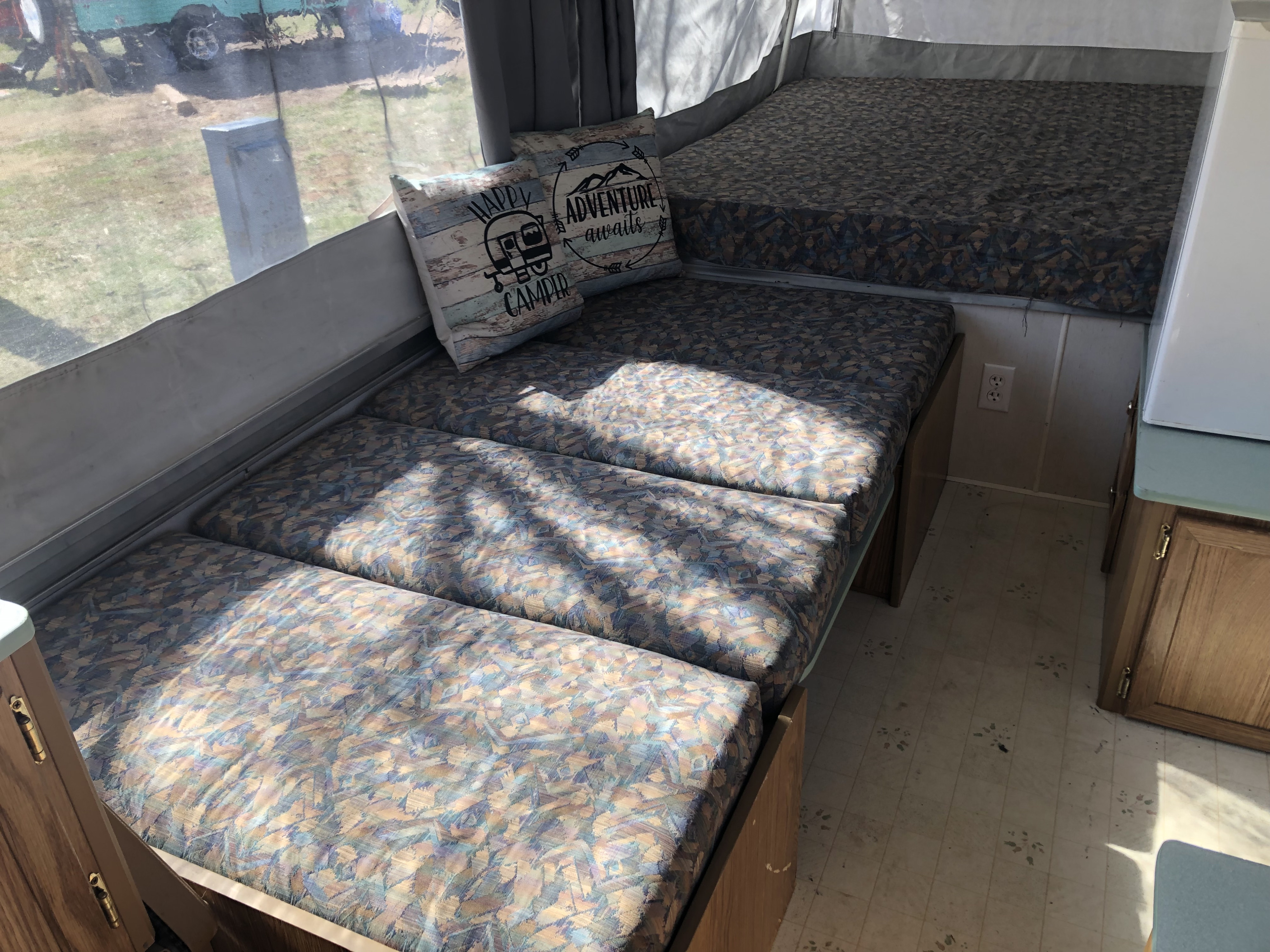 Interior of an RV rental featuring patterned bench cushions, decorative 'Happy Camper' pillows, and a sunlit window view.. Coleman Chesapeake 1993