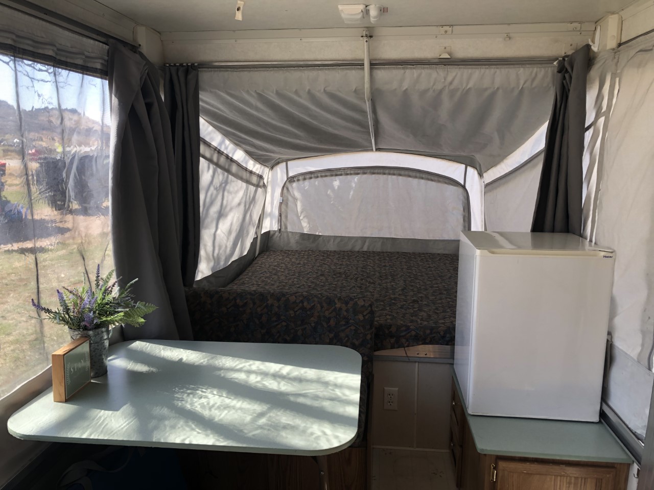 Cozy pop-up RV rental interior with a bed, dining table, mini-fridge, and sunlit outdoor views.. Coleman Chesapeake 1993
