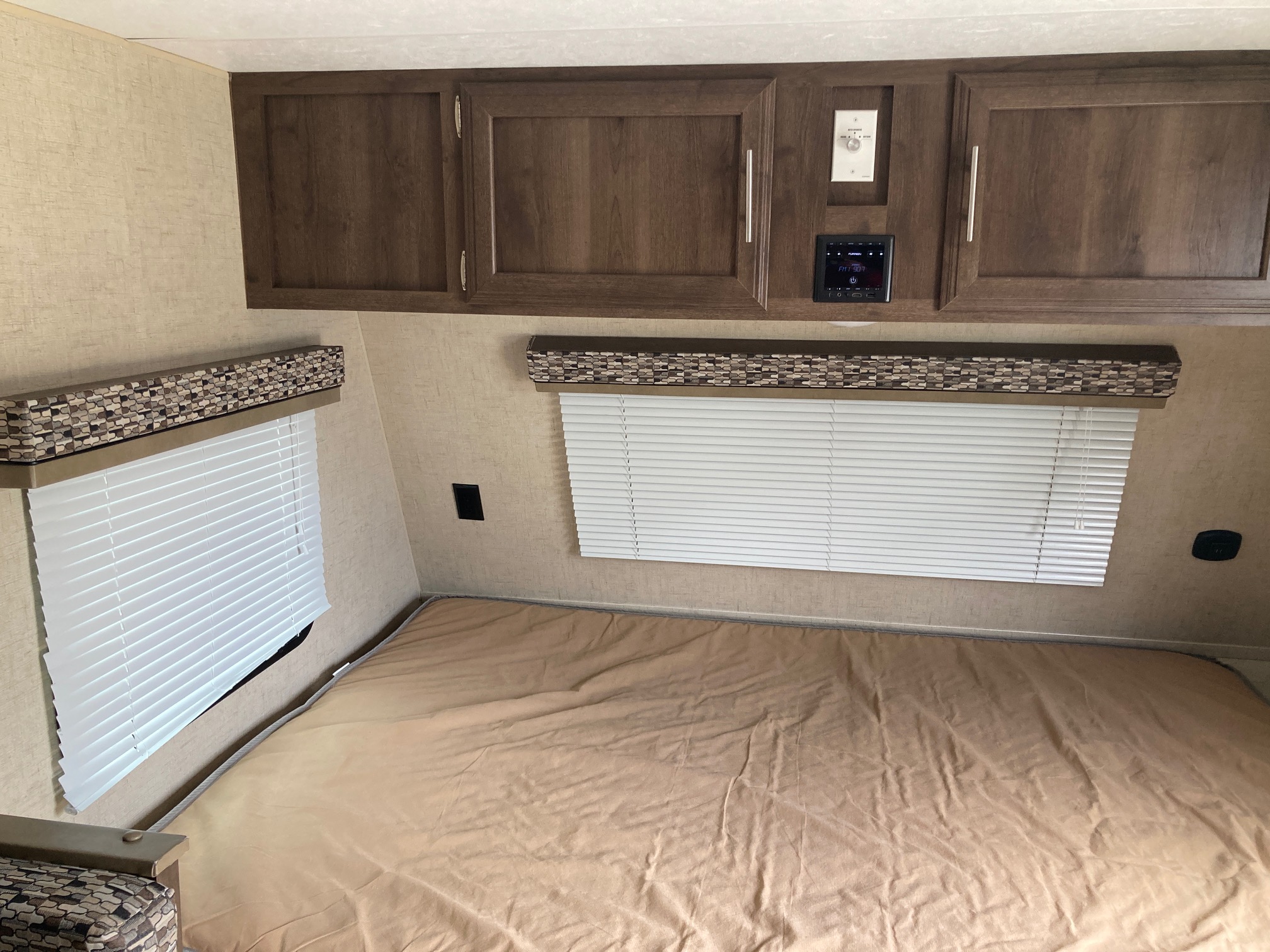 Inside an RV rental, a cozy bedroom with a tan bed, wood overhead cabinets, and windows with white blinds.. KZ Escape 2017