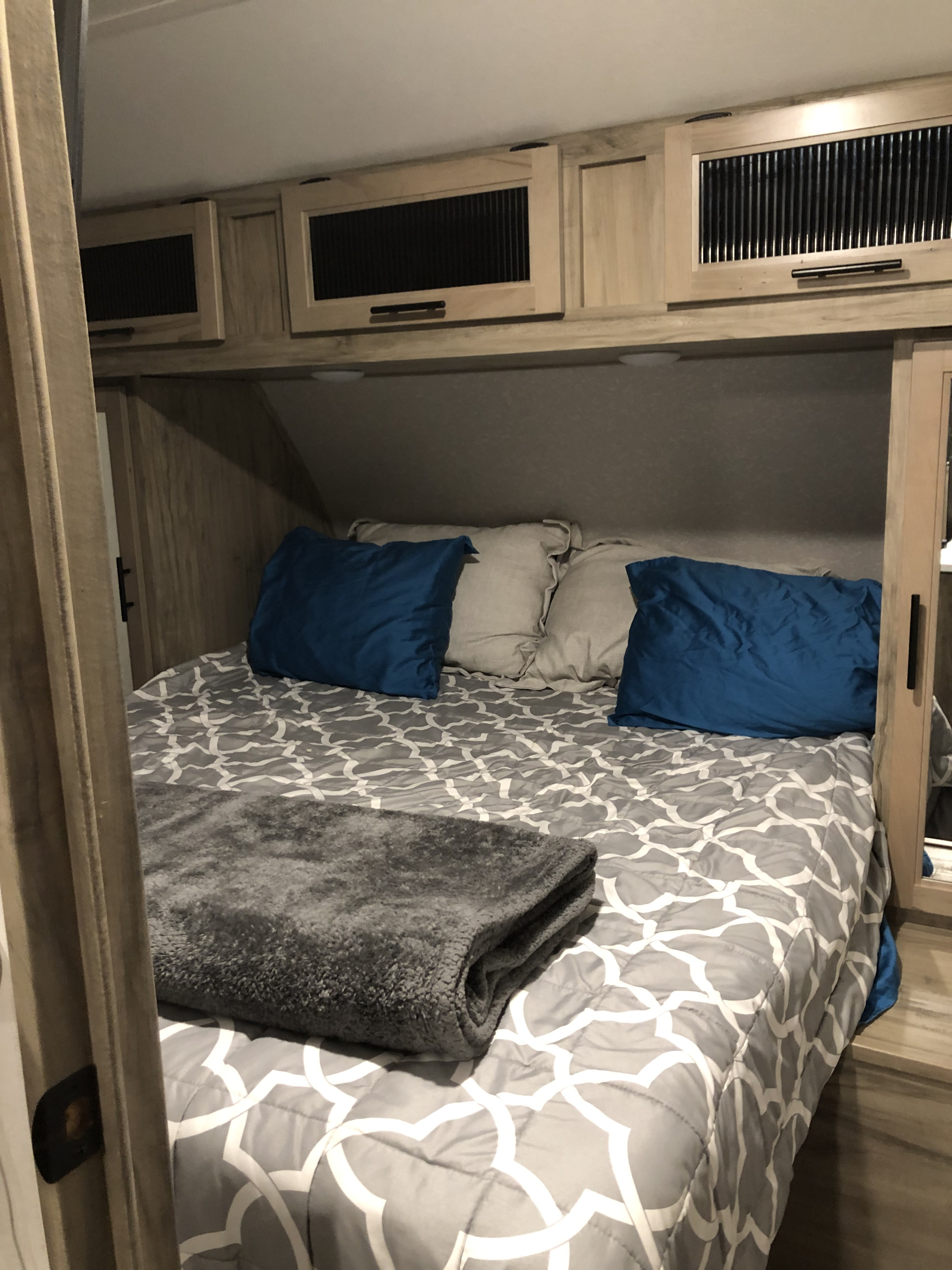 Cozy RV rental bedroom featuring a patterned bed with blue pillows and light wooden storage cabinets overhead.. Forest River Cherokee Alpha Wolf 2021
