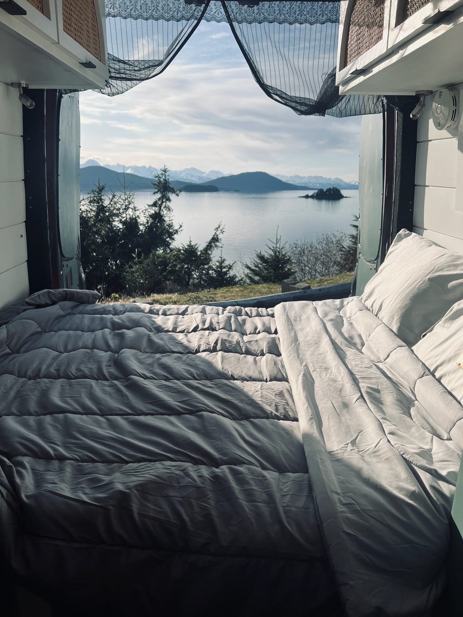 An RV rental bedroom with a view! Cozy bed overlooks a serene lake, evergreen trees, and snowy mountains. Perfect travel.. Freightliner Sprinter 2011
