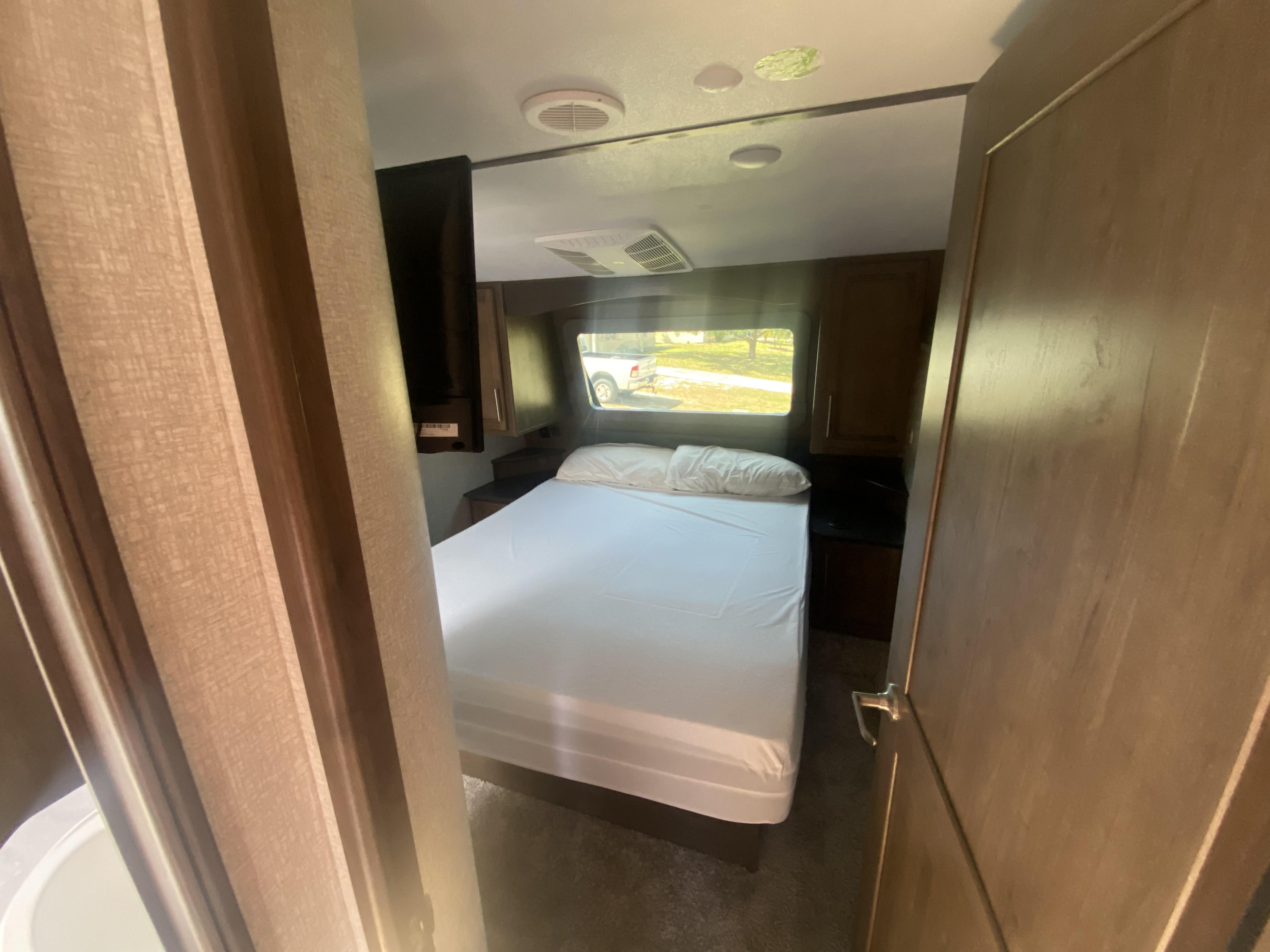 Peek into an RV rental bedroom with a white-sheeted bed, ceiling AC, mounted TV, and a window offering a view outside.. Keystone RV Cougar 2021