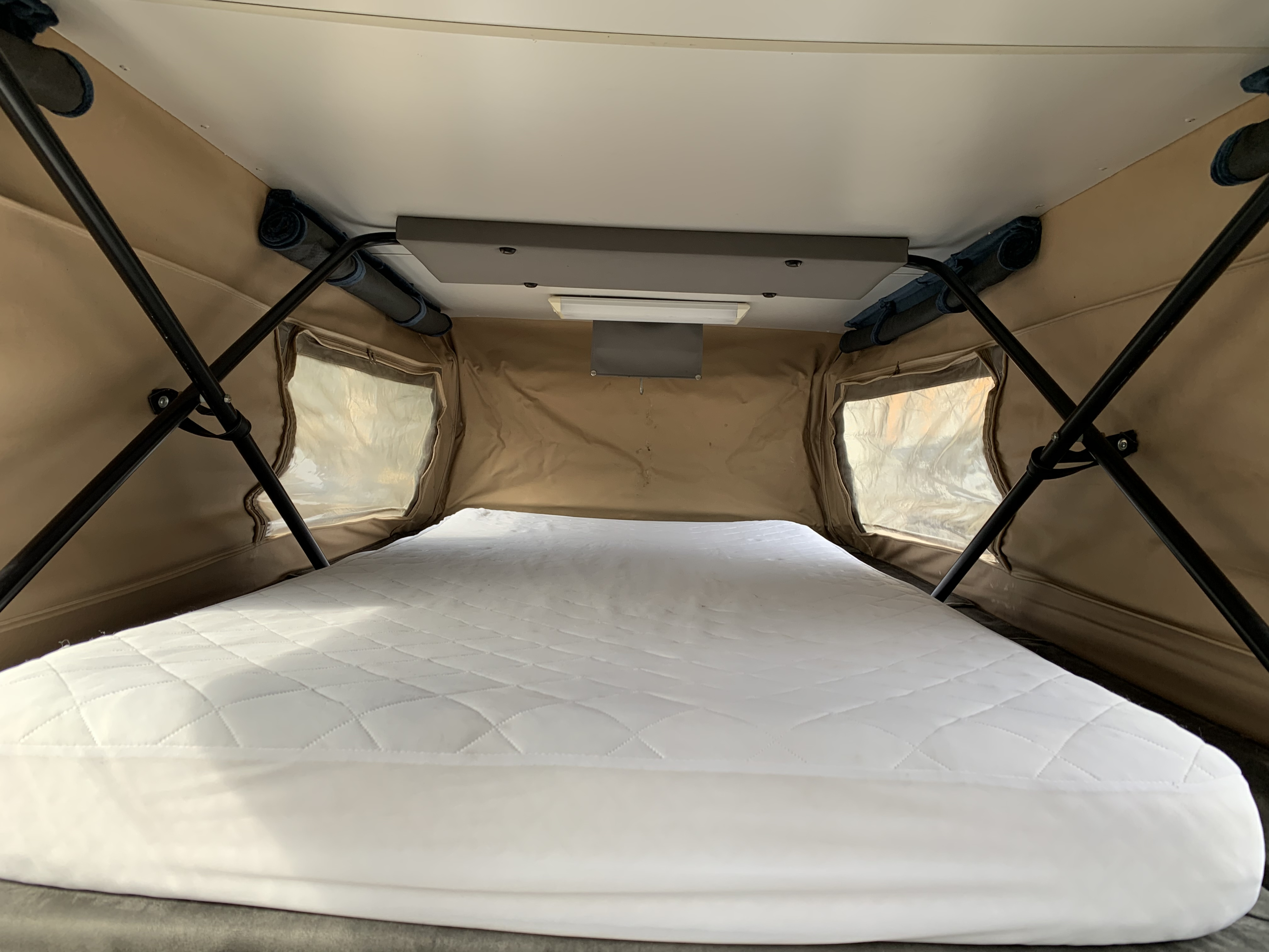Inside an RV rental pop-top camper: A comfortable sleeping area with a white quilted mattress, beige fabric walls, and clear windows.. Sportsmobile Chevy 3500 2001