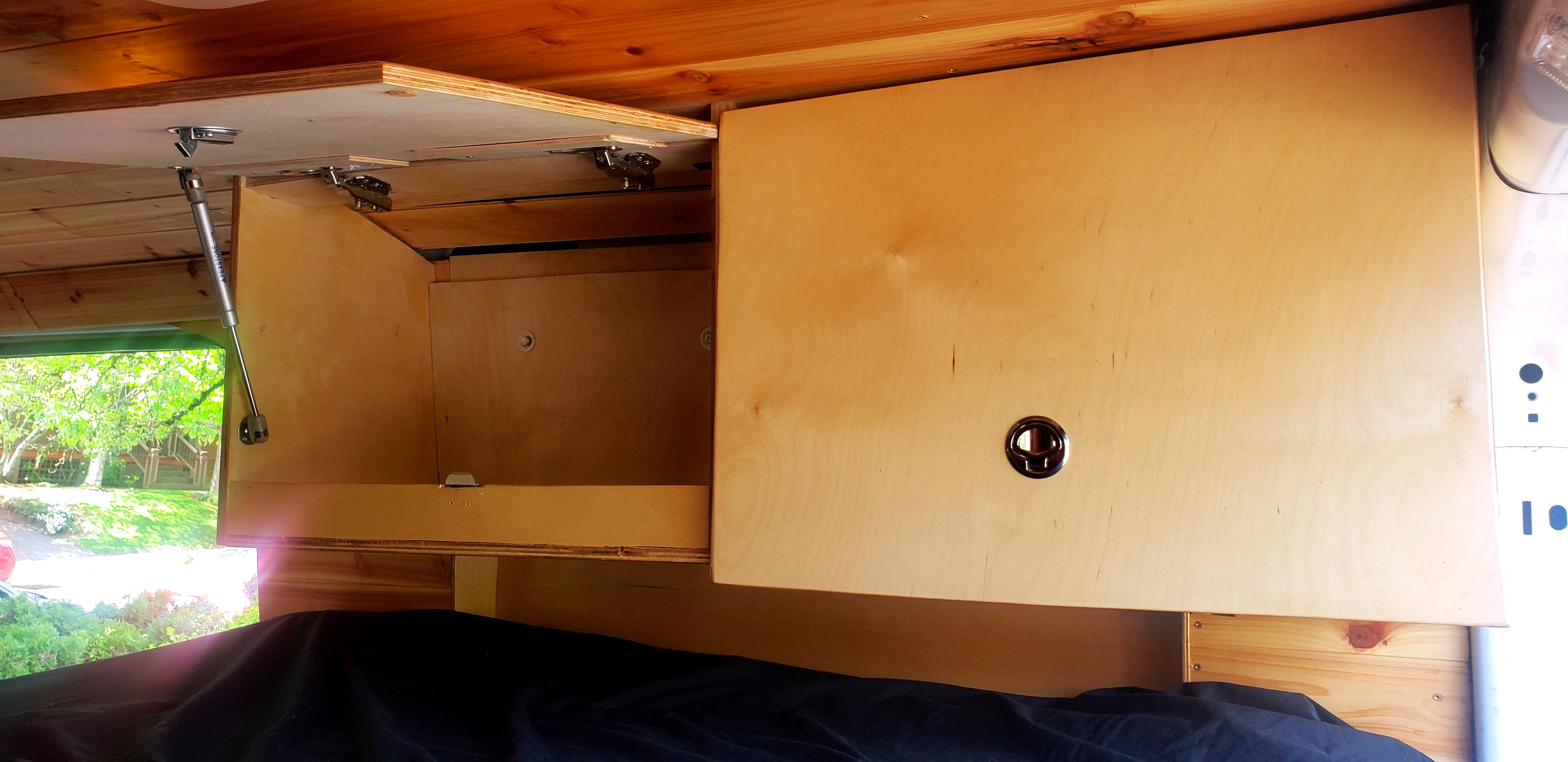 Wood overhead cabinets inside an RV rental. One open with a gas strut, one closed. Glimpse of trees outside.. Ford Transit 2018