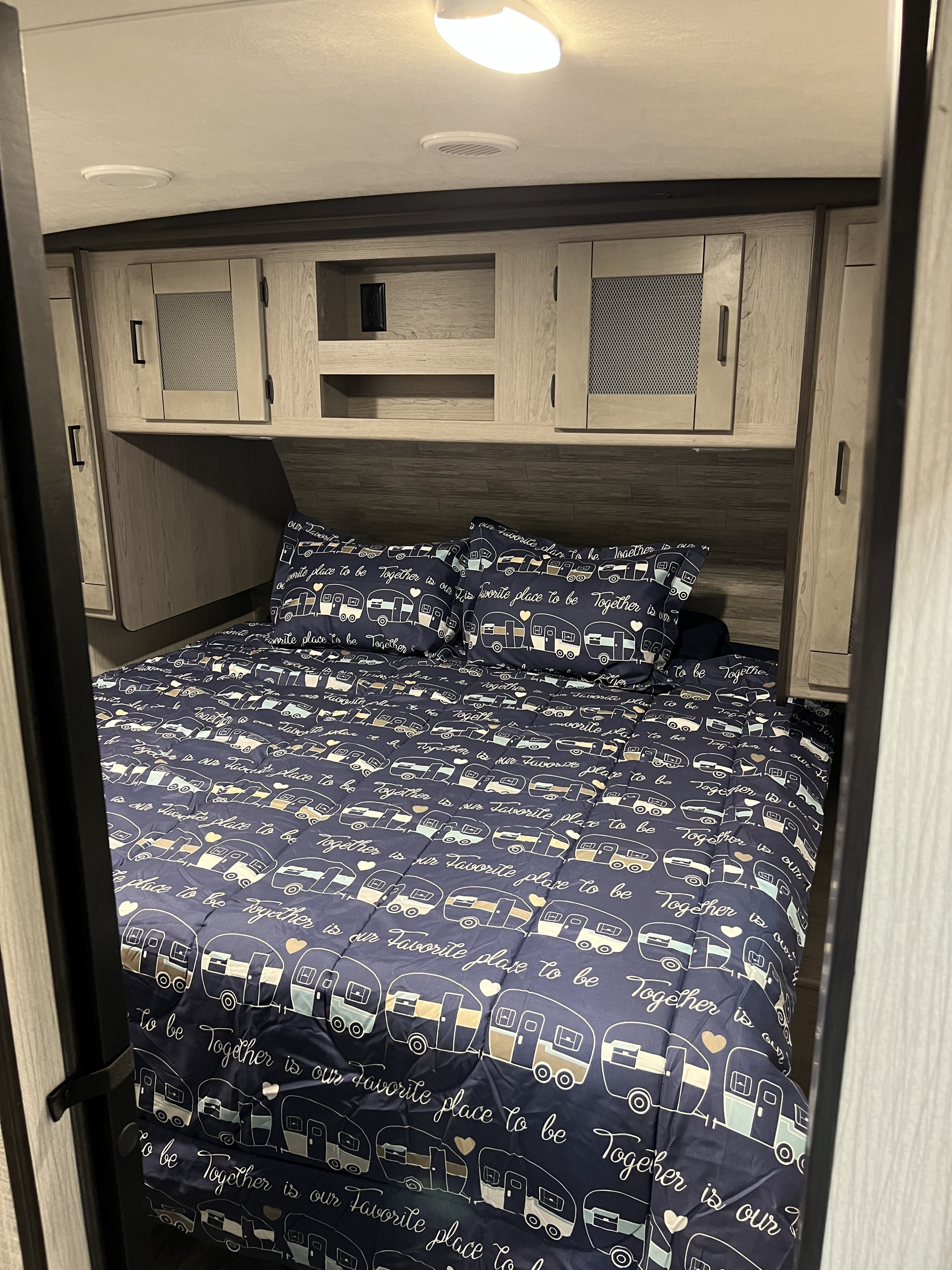 Cozy RV rental bedroom. Features a bed with navy camper-themed bedding, wood overhead cabinets, and ceiling light/vent.. Heartland RVs Mallard 2021