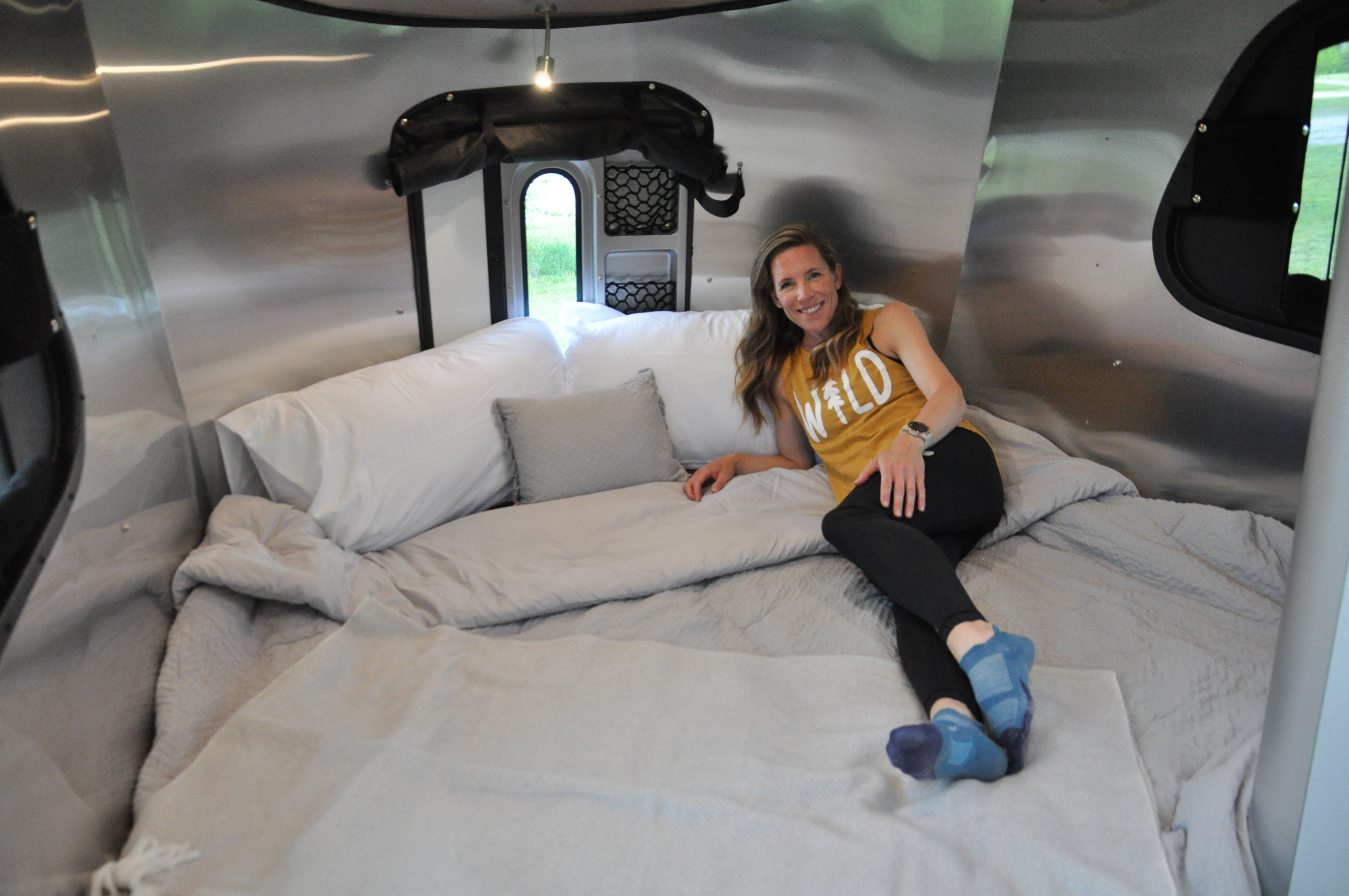 Woman smiles lounging in a cozy RV rental bed, with sleek metallic interior and outdoor views. Perfect for travel.. Airstream Basecamp 2022