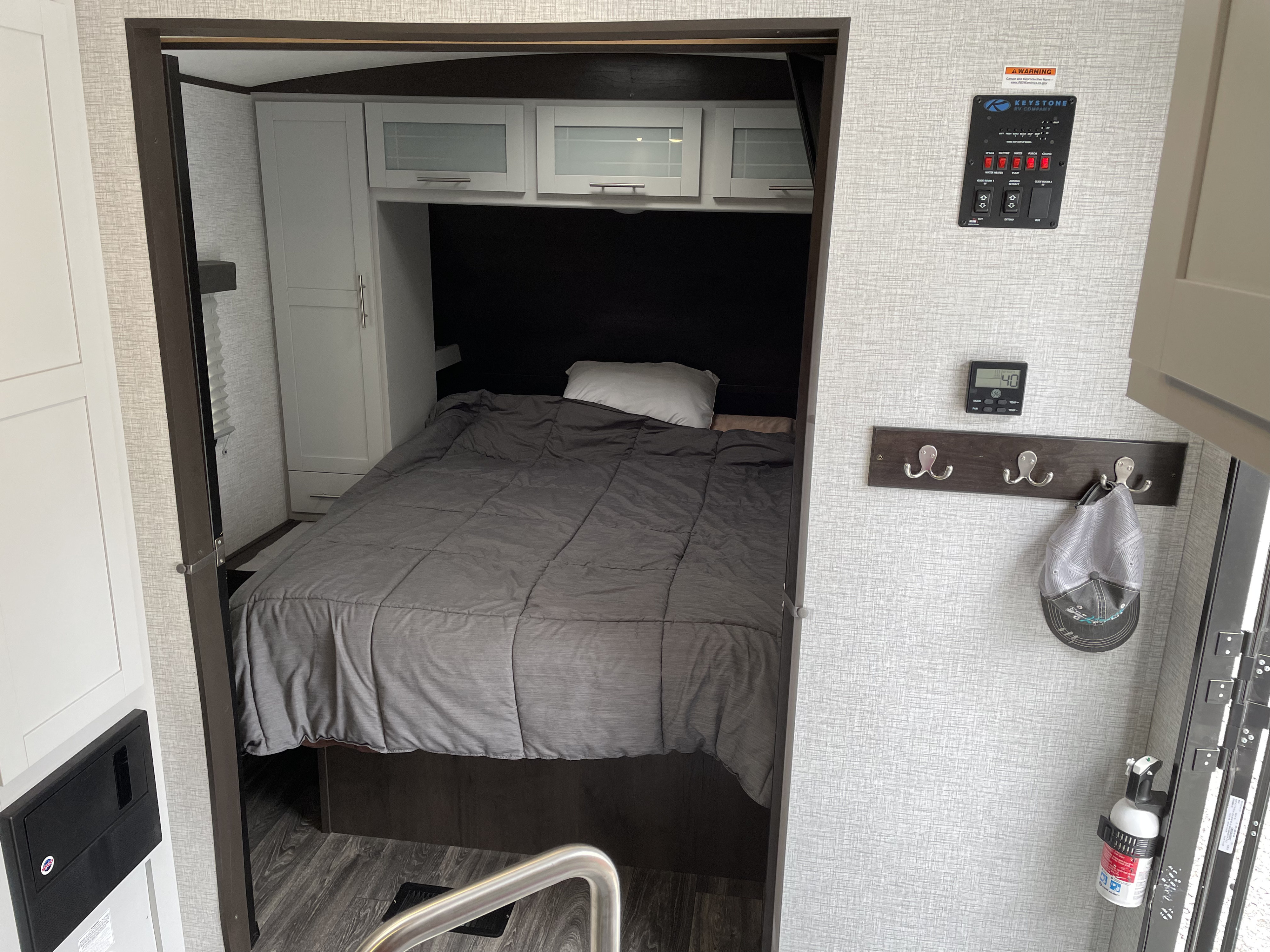 Step into this clean RV rental bedroom, featuring a comfortable bed, overhead cabinets, and essential control panel. Ideal for travel.. Keystone RV Bullet Ultra Lite 2021