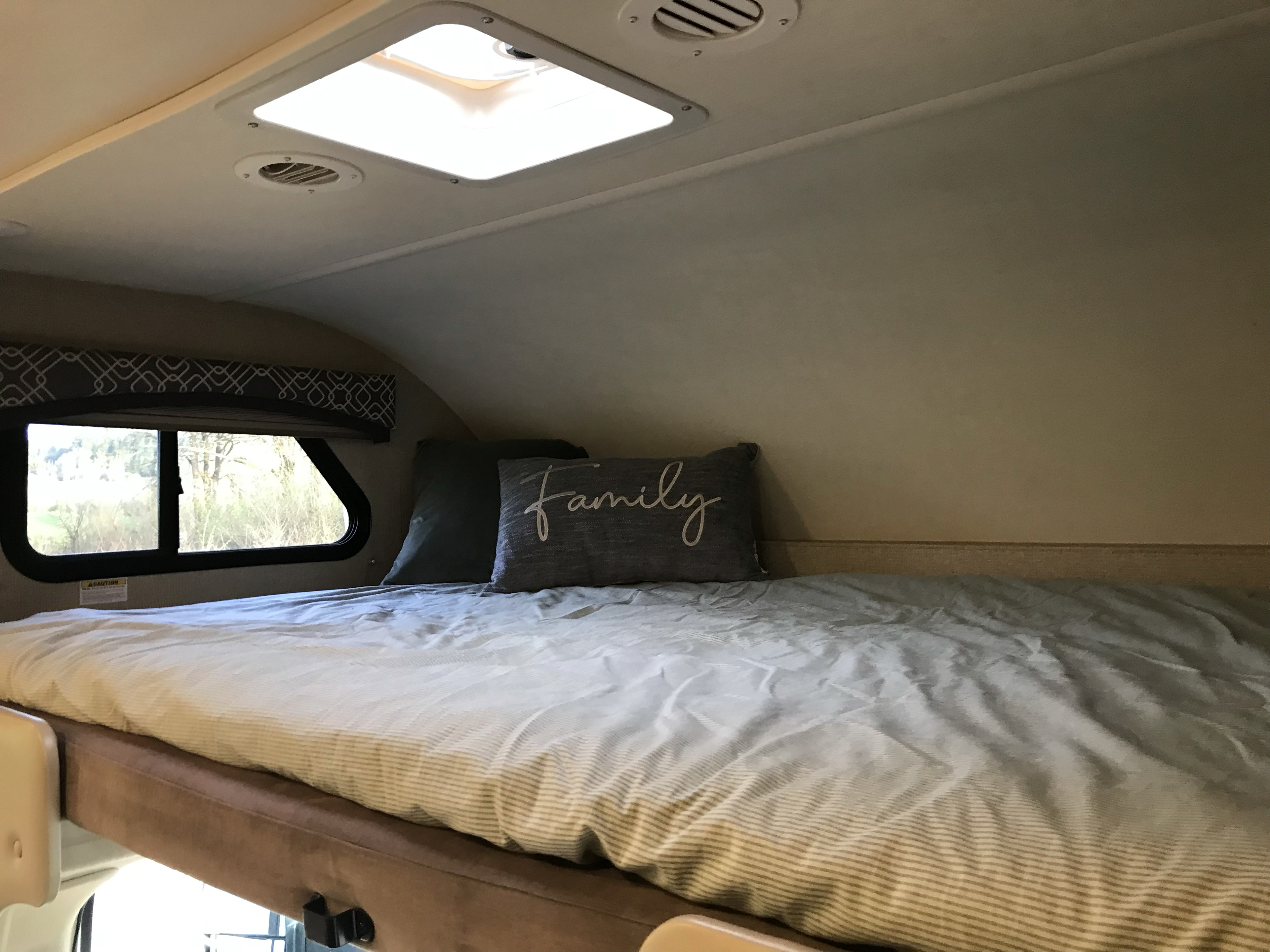 RV rental interior: a comfy cab-over bunk bed with striped bedding, "Family" pillow, window, and ceiling vents.. Thor Chateau 2019