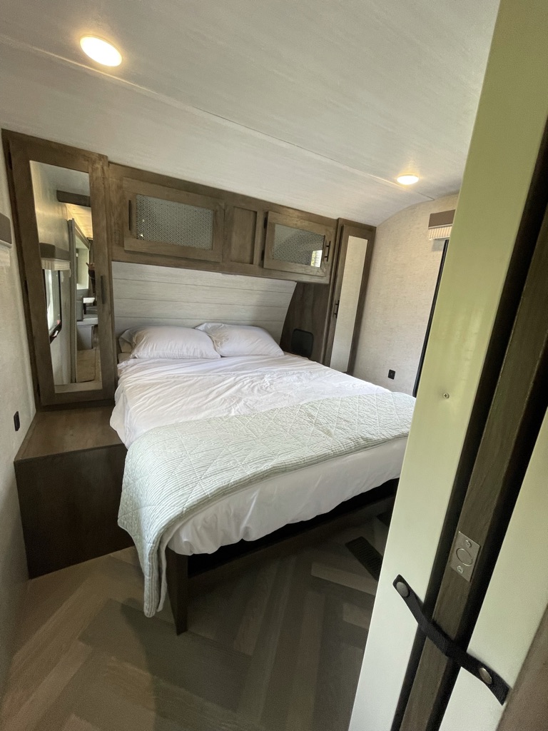 An RV rental bedroom with a neatly made queen bed, white and green bedding, overhead storage, and modern decor.. Forest River Other 2021