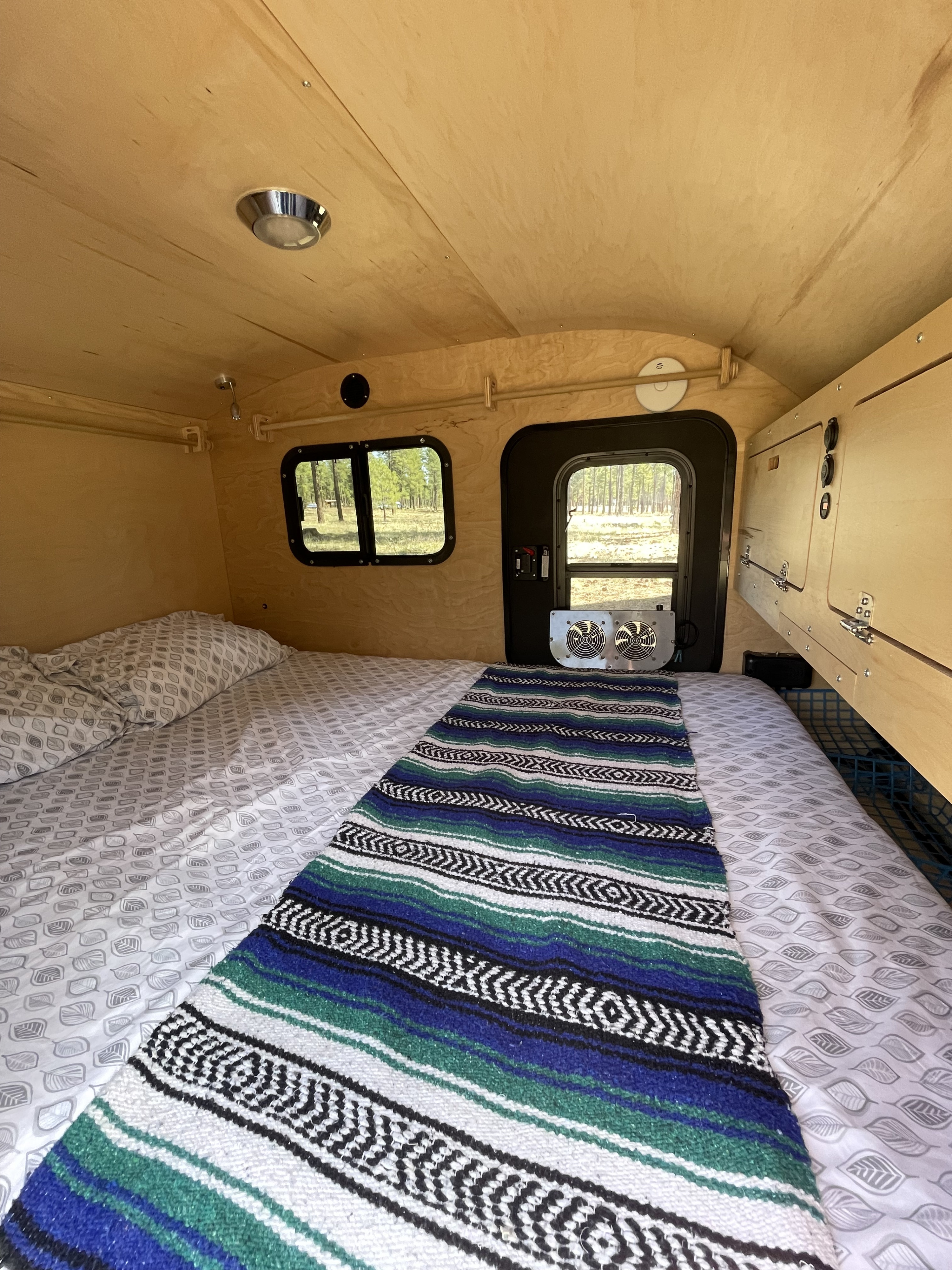 Inside an RV rental: a cozy wooden interior with a bed, patterned bedding, and colorful blanket. Windows offer forest views.. TC Teardrop Overland 2021