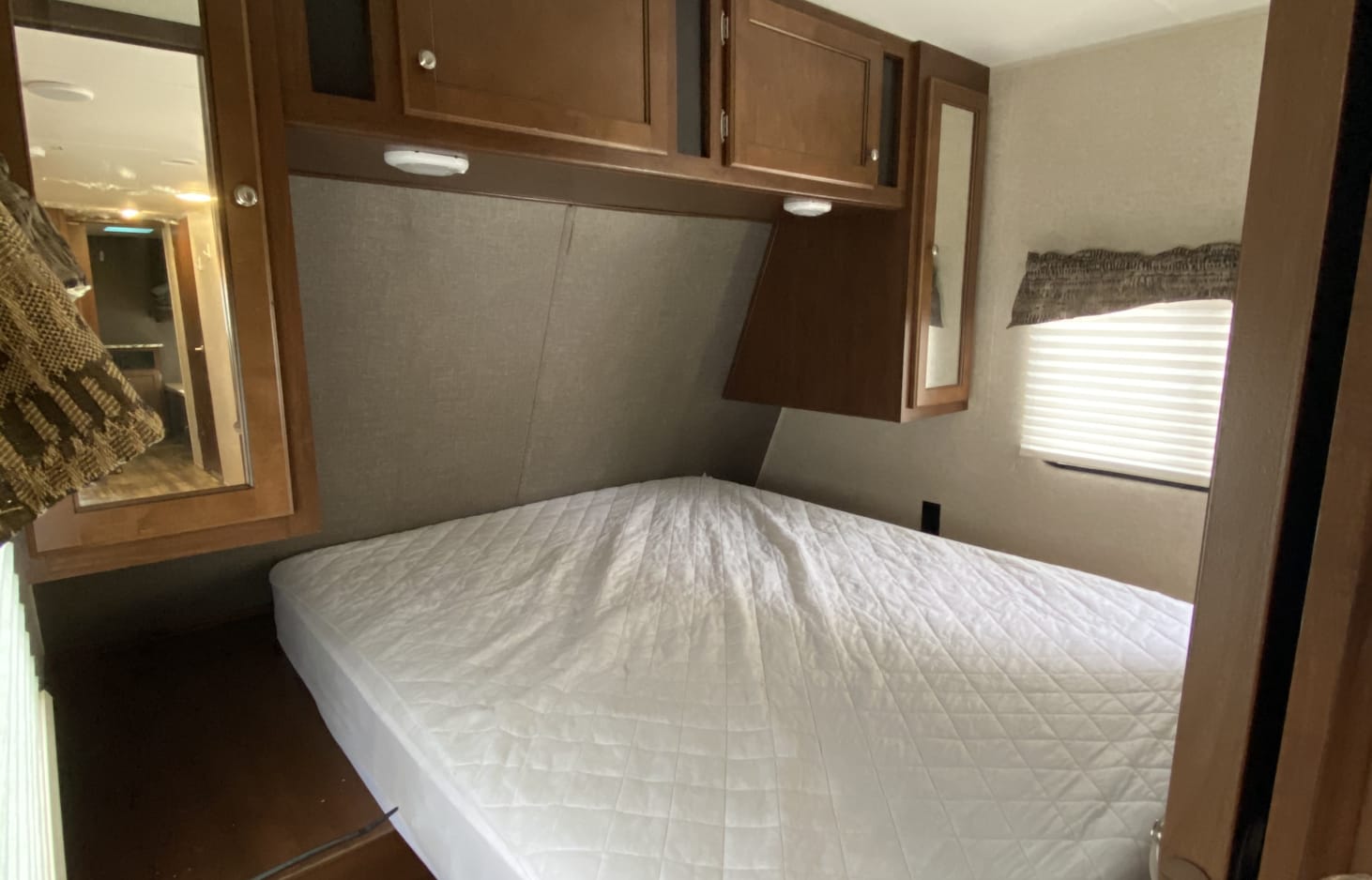 An RV rental bedroom with a neat bed, wooden overhead cabinets, a mirror, and a window with blinds.. Heartland Trail Runner 2017