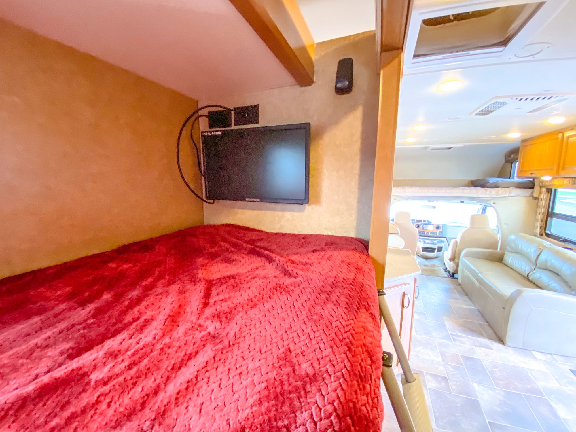 RV rental interior: A bunk bed with a red blanket and mounted TV is in the foreground, with the spacious living area visible behind.. Thor Chateau 2016