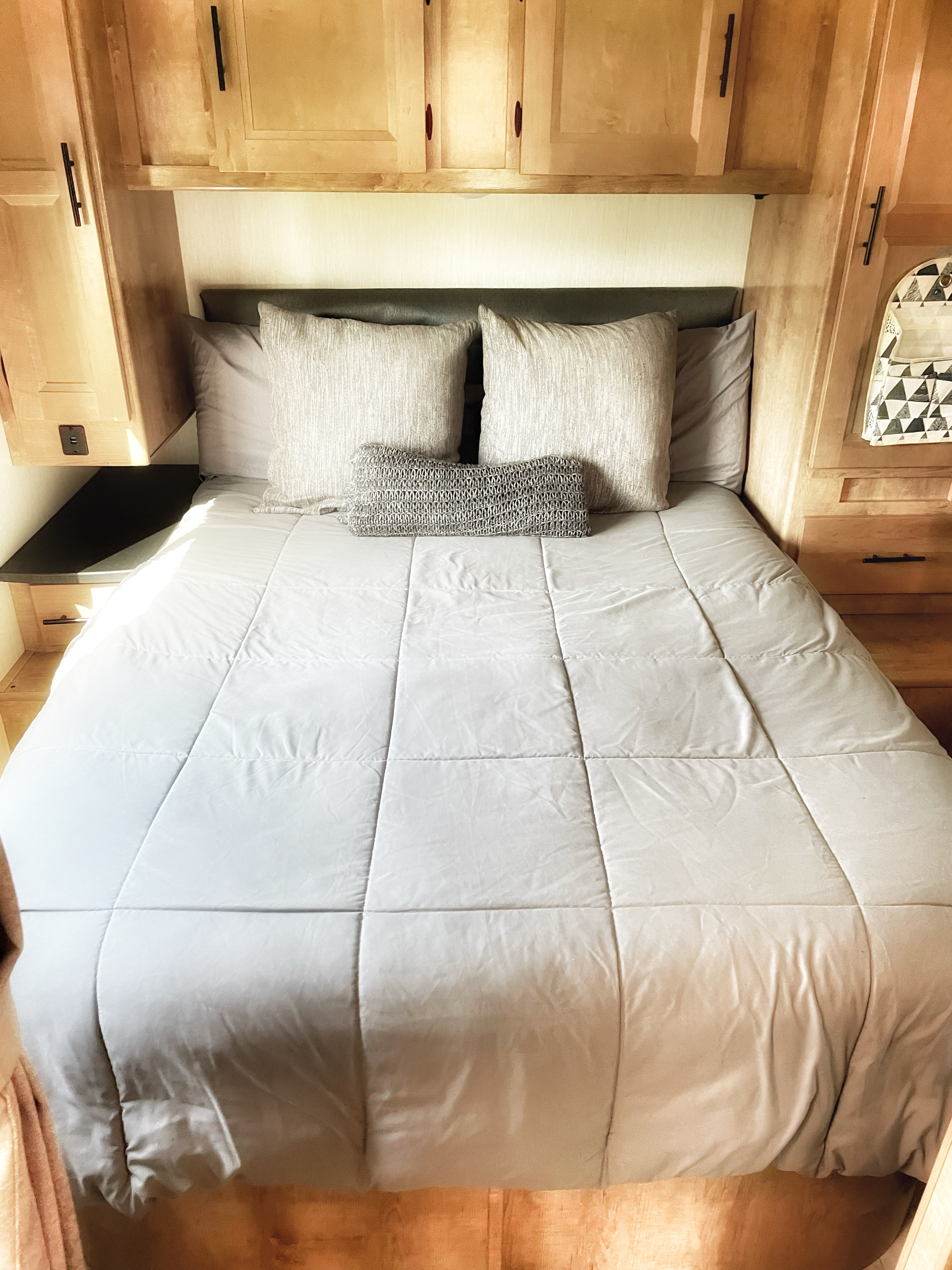 Cozy RV rental bedroom featuring a comfortable bed with grey bedding, light wood cabinetry, and integrated charging options.. Other Other 2021