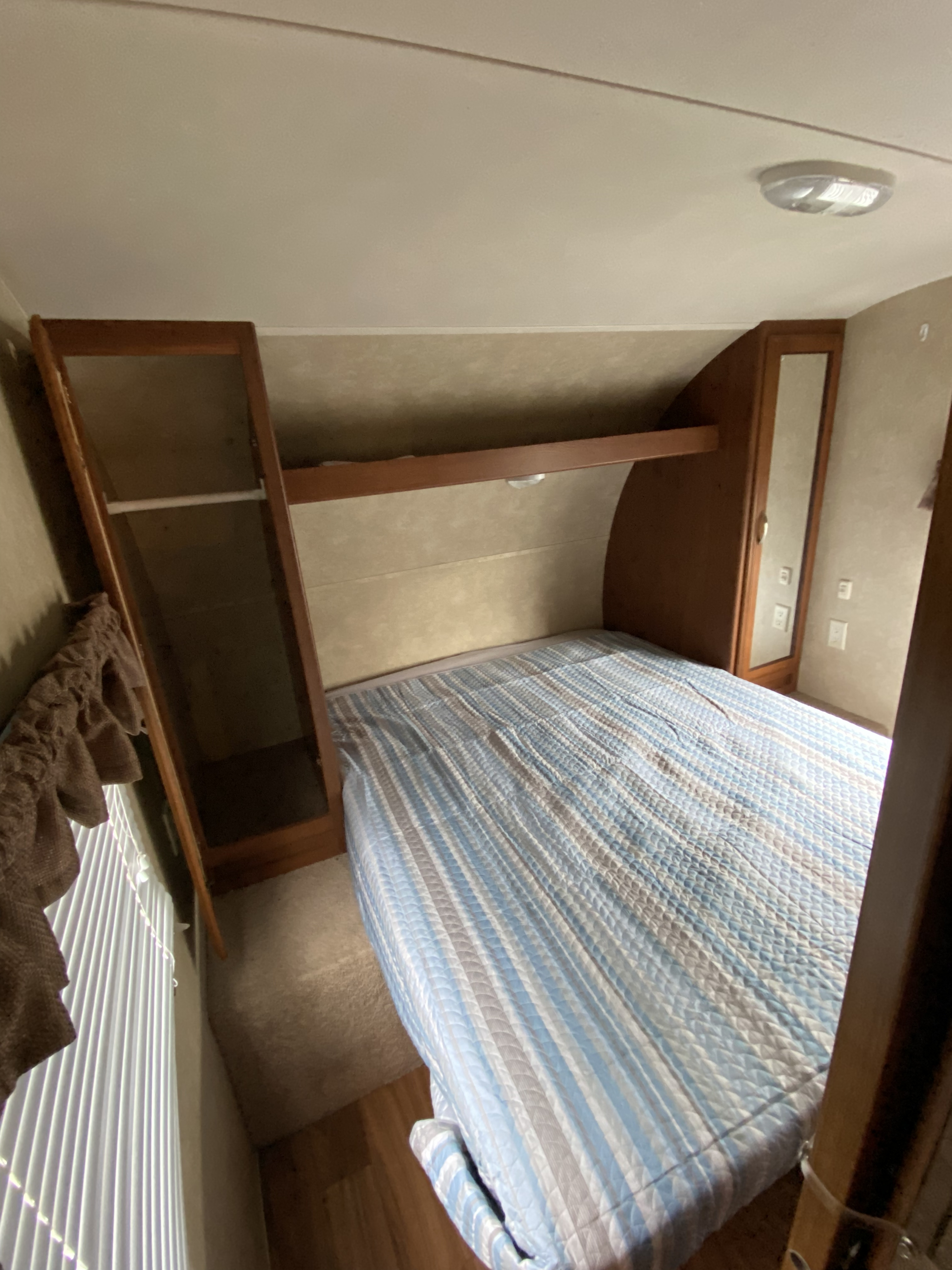 Inside an RV rental: a compact bedroom with a blue striped bed, built-in wood cabinets, shelf, and mirror, plus a window.. Forest River Wildwood X-Lite 2014