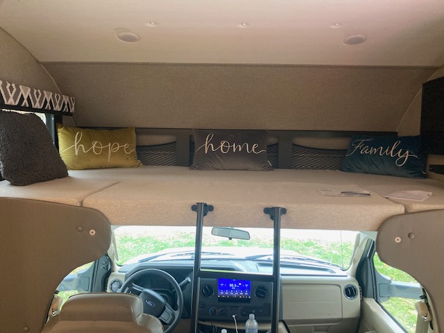 Inside an RV rental: a spacious cab-over bed adorned with "hope," "home," and "family" pillows, overlooking the cockpit.. Jayco Greyhawk 2021