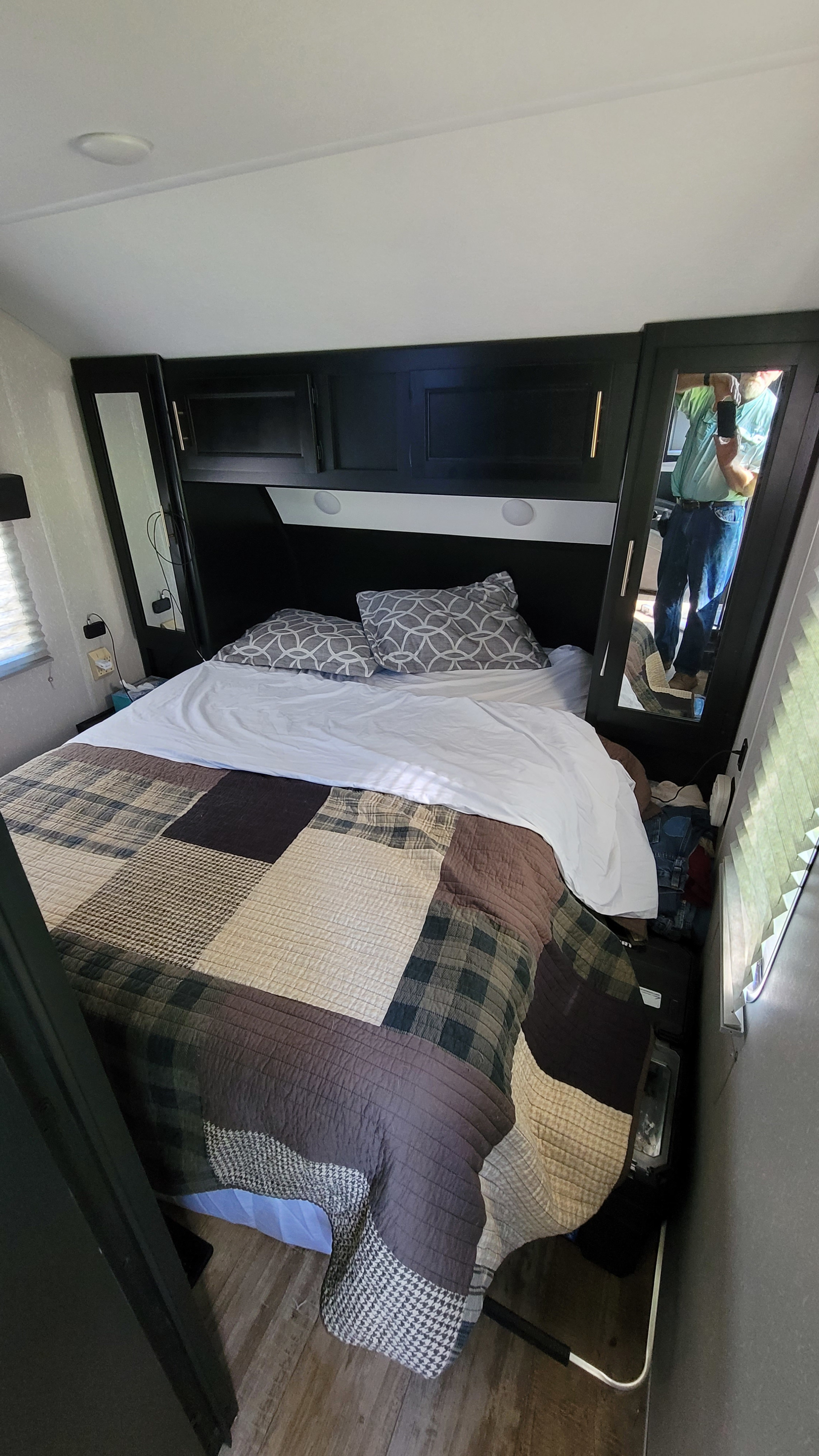 Cozy bedroom inside an RV rental, with a plaid quilt, dark headboard, and mirrored cabinets. A person is visible in the reflection.. Forest River Cherokee 2021
