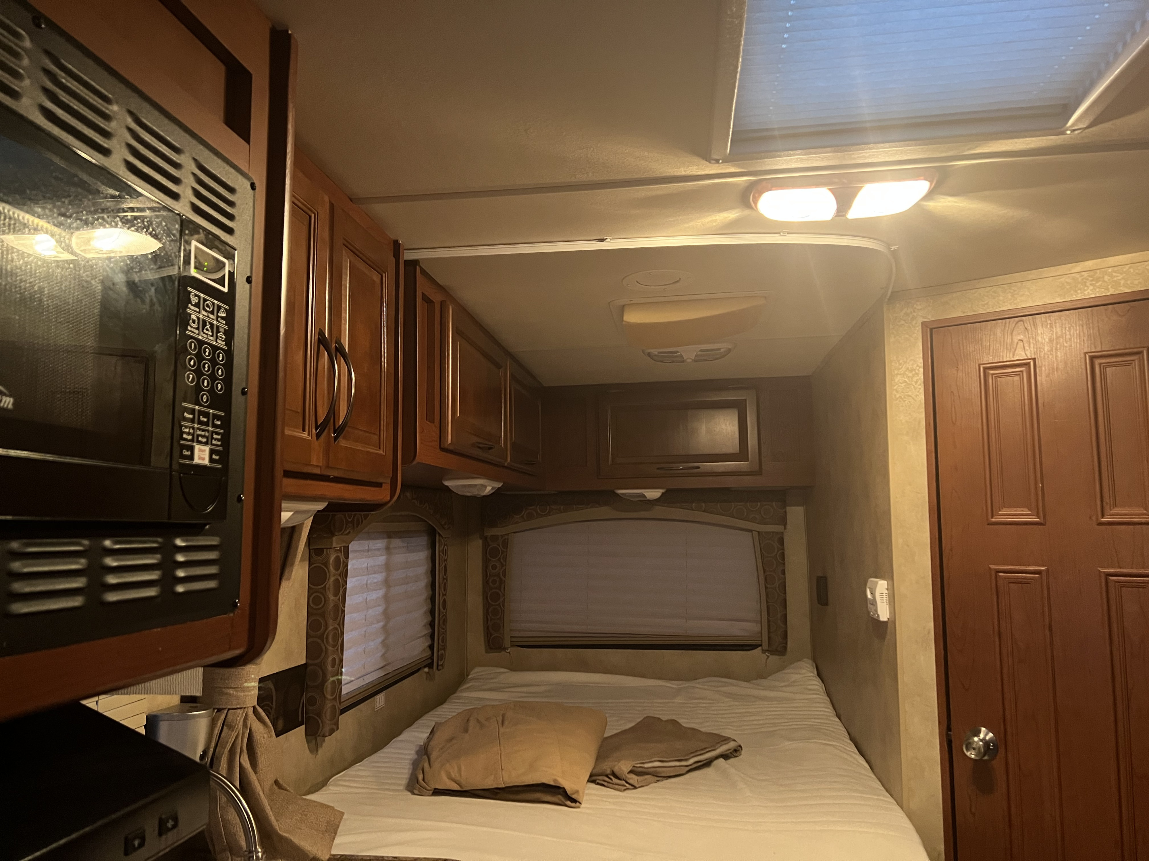 Inside a cozy RV rental: a bedroom and kitchenette area with a microwave, bed, wooden cabinets, windows, and ceiling lights.. Thor Chateau 2012
