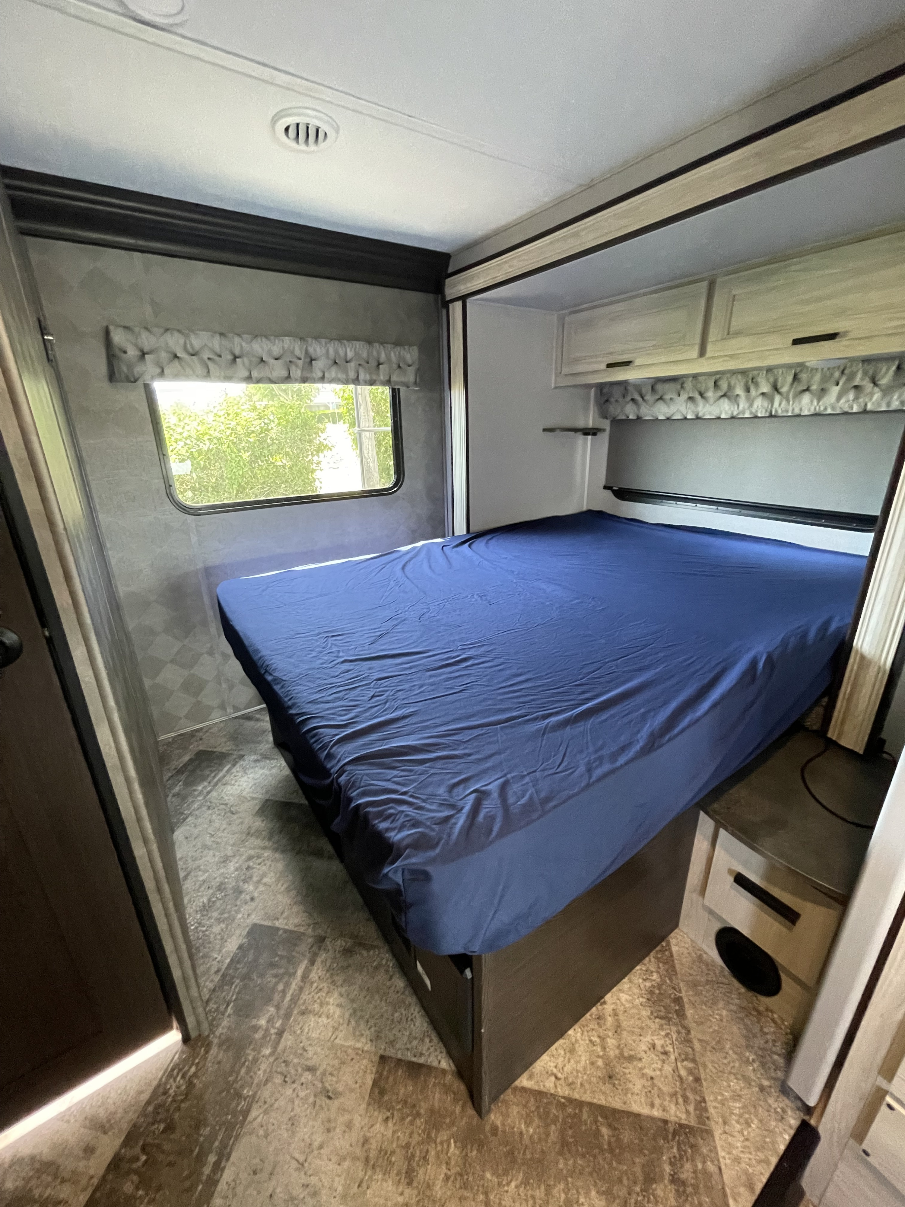 An RV rental bedroom showcasing a blue bed, light wood cabinets, and a window with a green view. Functional and inviting.. Forest River Sunseeker 2021