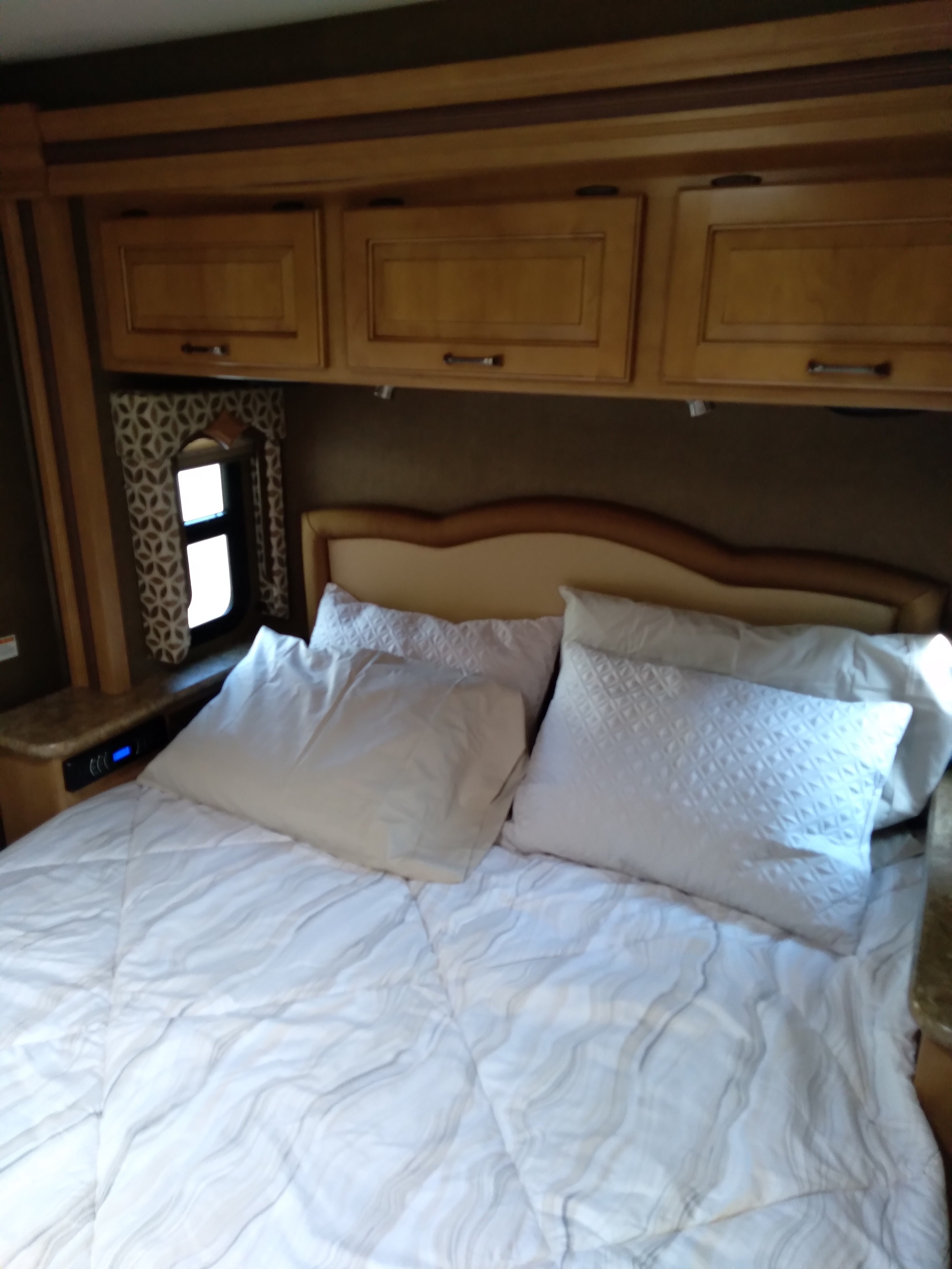 Cozy RV rental bedroom with a large bed, white bedding, wooden overhead storage, and a small side window.. Thor Hurricane 2016